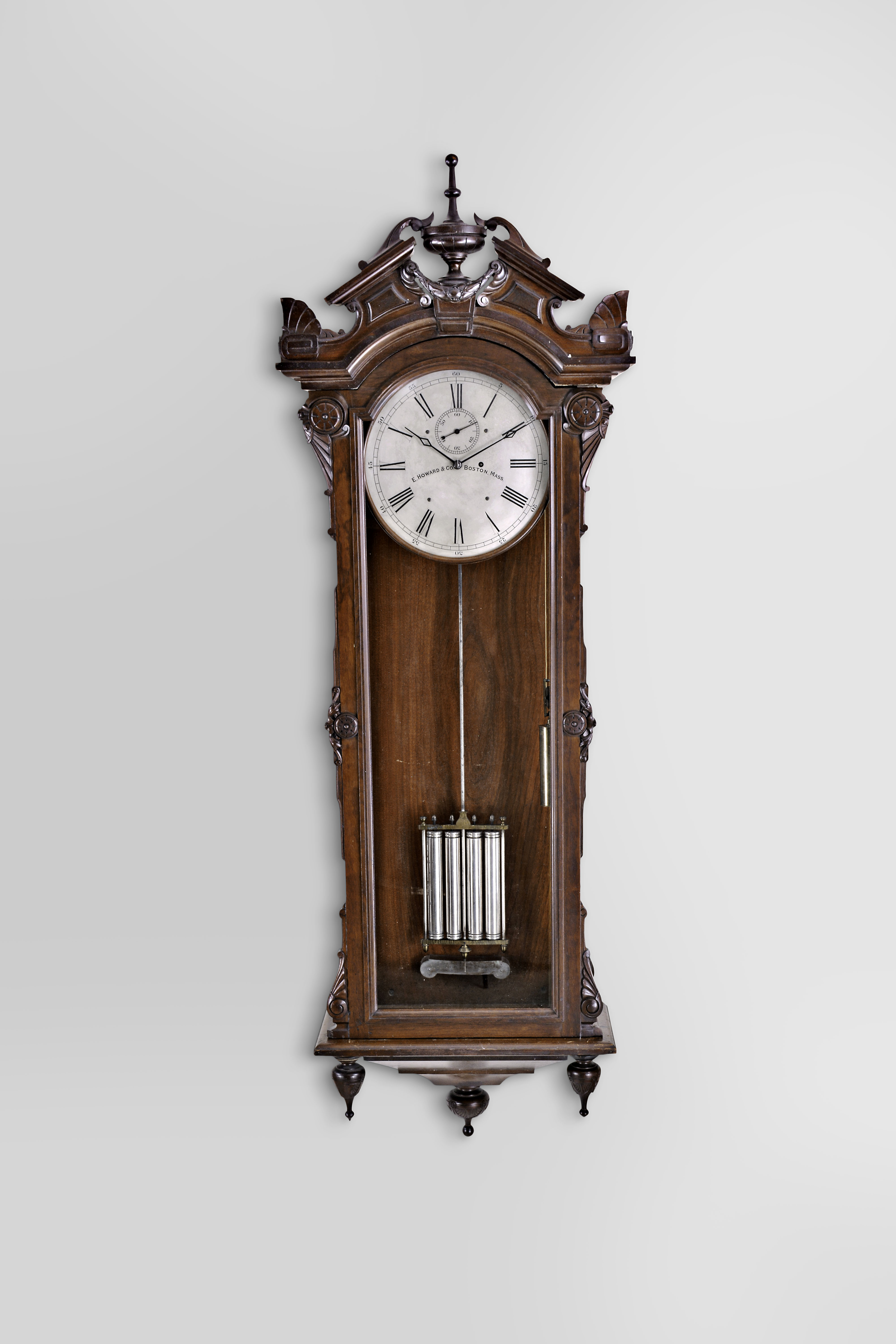 E. Howard &amp; Co., Boston, "No. 60" hanging regulator, the renaissance revival style case with glazed door and sides, the door with applied, stylized Grecian foliate carvings, and simple eight lobe roundels, base with two turned, carved drops flanking a tapered, stepped bracket terminating in a large, shallow cavetto molding with turned and carved drop, the top with arched cornice molding surmounted by a broken pitched pediment with central urn and laurel festoon, and the corners with stylized palmette form acroterion angularium above stylized, flattened volutes, silvered dial signed "E. Howard &amp; Co. Boston, Mass.", blued steel hands, damascened brass week going movement with jewelled pallets, mercury pendulum with four nickel plated steel jars, brass frame, and nickel plated rod, silvered beat scale, brass clad weight, and heavy cast iron bracket attached to the case back, supporting both the pendulum and the movement — Antique Clocks Price Guide