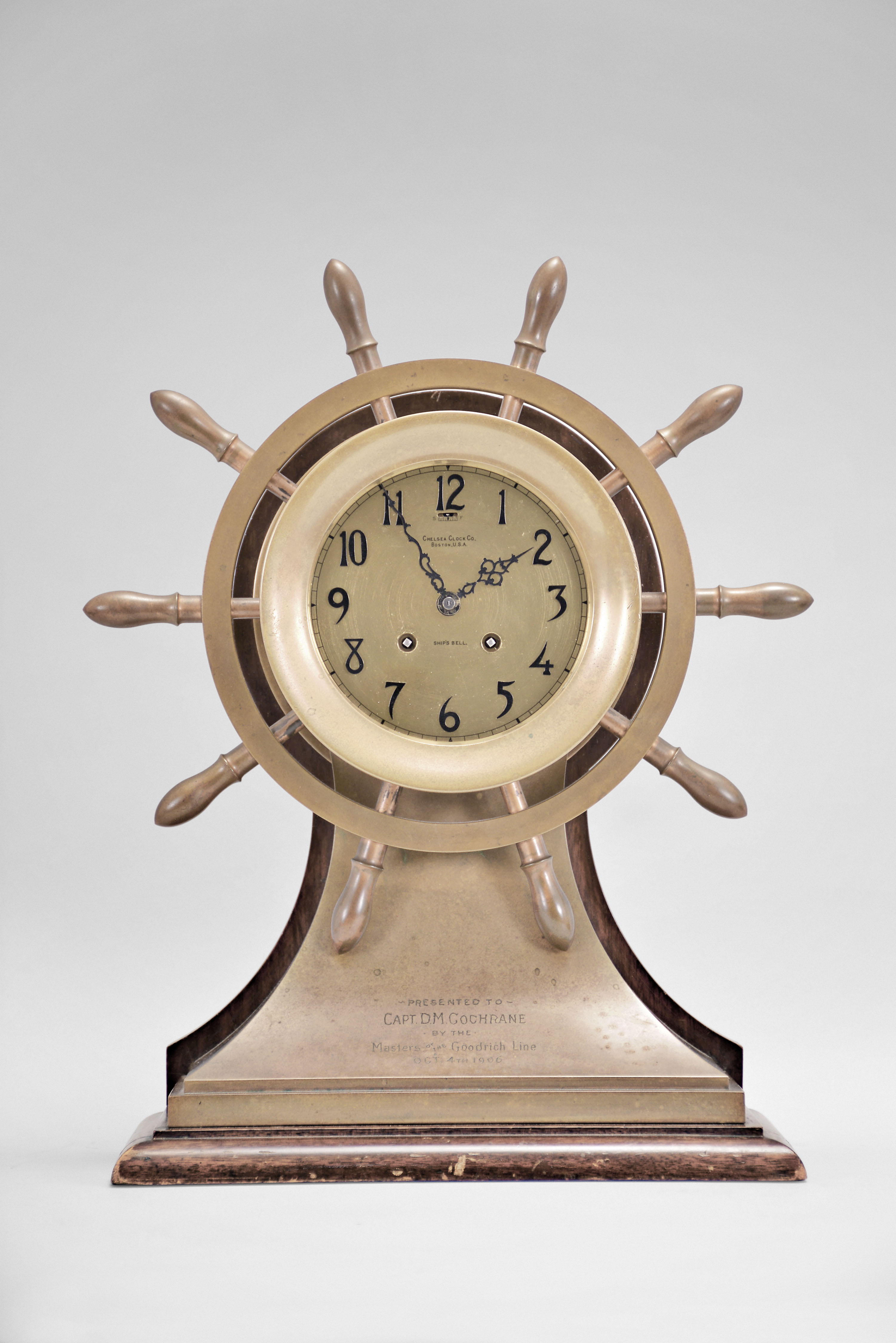 Chelsea Clock Co., Boston, Mass., "Mariner" shelf clock with six inch dial, polished brass case in form of yacht's wheel engraved "PRESENTED TO CAPT. D.M. COCHRAN BY THE Masters of the Goodrich Line Oct. 4 1906" mounted on mahogany stand, signed Arabic numeral silvered special dial with applied numerals also marked "Ships Bell", blued steel hands, 8 day, time and strike movement with lever escapement, serial #15227 including Chelsea marked key — Antique Clocks Price Guide