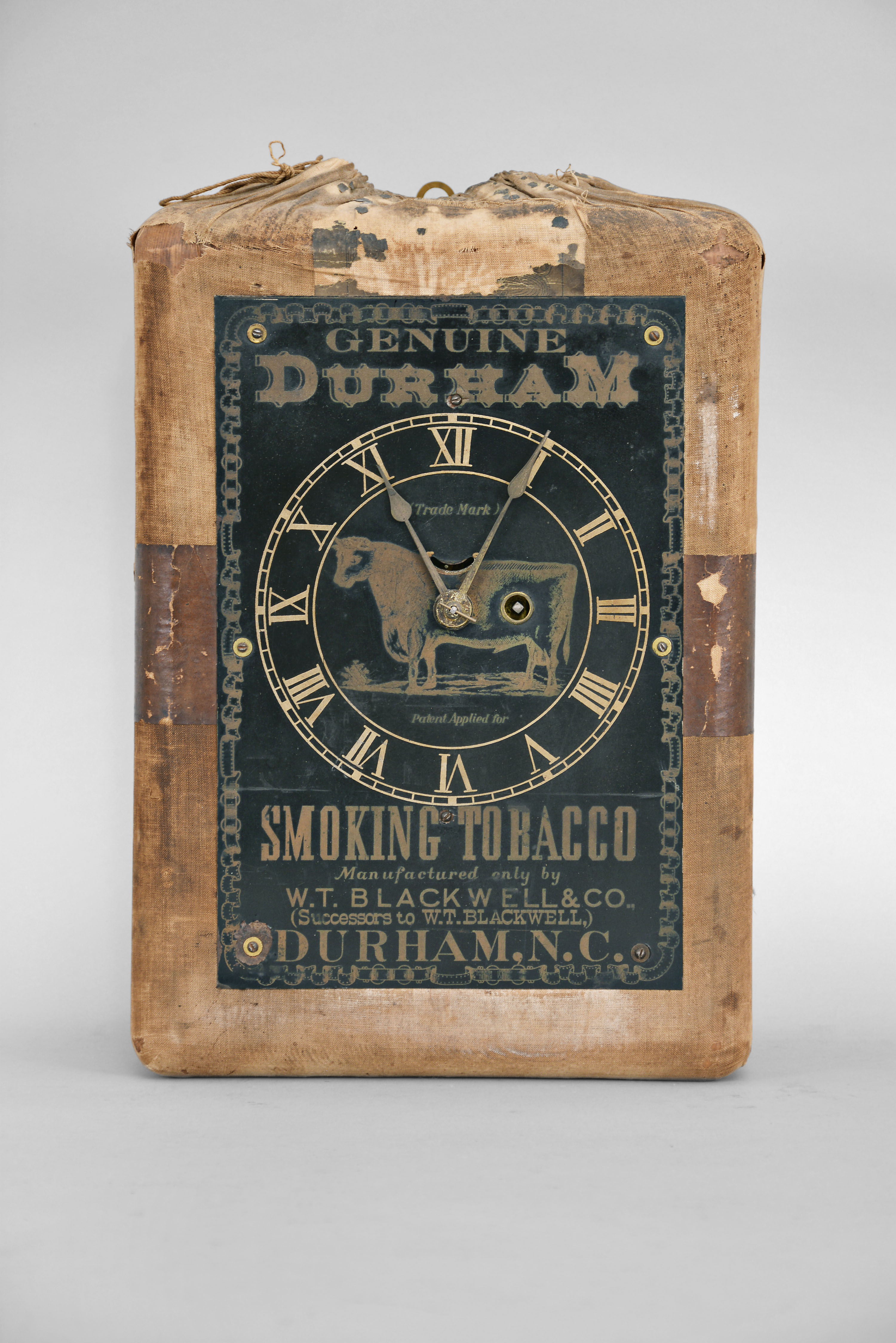 Ansonia Clock Co., New York, for Bull Durham Tobacco, advertising clock, 30 hour, spring driven balance wheel movement in a wood case wrapped in cloth to resemble a bail of tobacco, black steel dial with advertising in gold print reading in part "Genuine Durham Tobacco / Durham, N.C.", gold colored spade hands. — Antique Clocks Price Guide