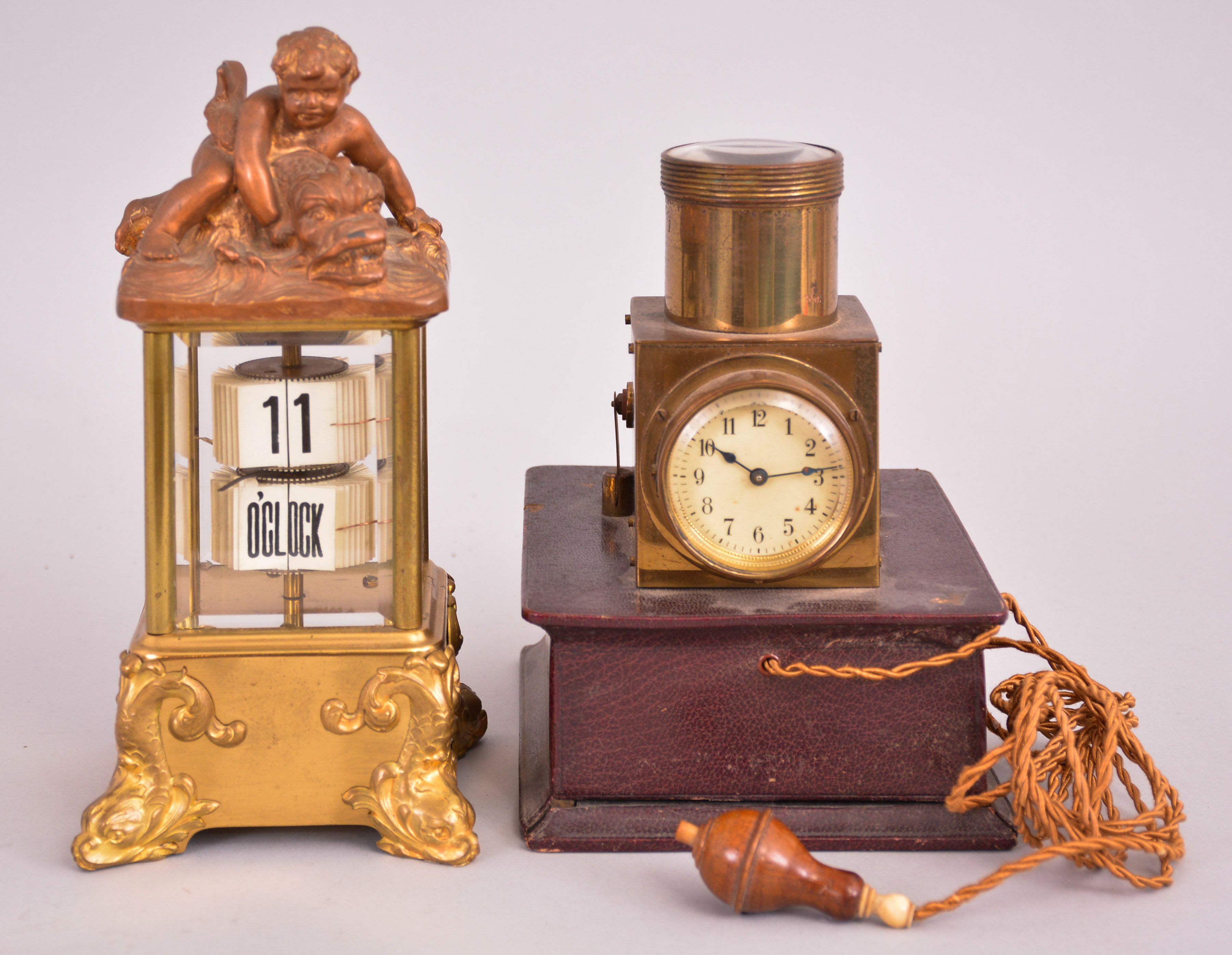 Clocks- 2 (Two): (1) Lenzkirch AG, Lenzkirch, Germany, digital flip card clock, spring driven balance wheel movement in a glazed case with dolphin-form feet, surmounted by a child wresting a sea creature, c1900; (2) Projection clock, 30 hour, time and light projection, spring driven balance wheel movement in a brass case with battery compartment in the base and corded push-button switch, c1920. — Antique Clocks Price Guide