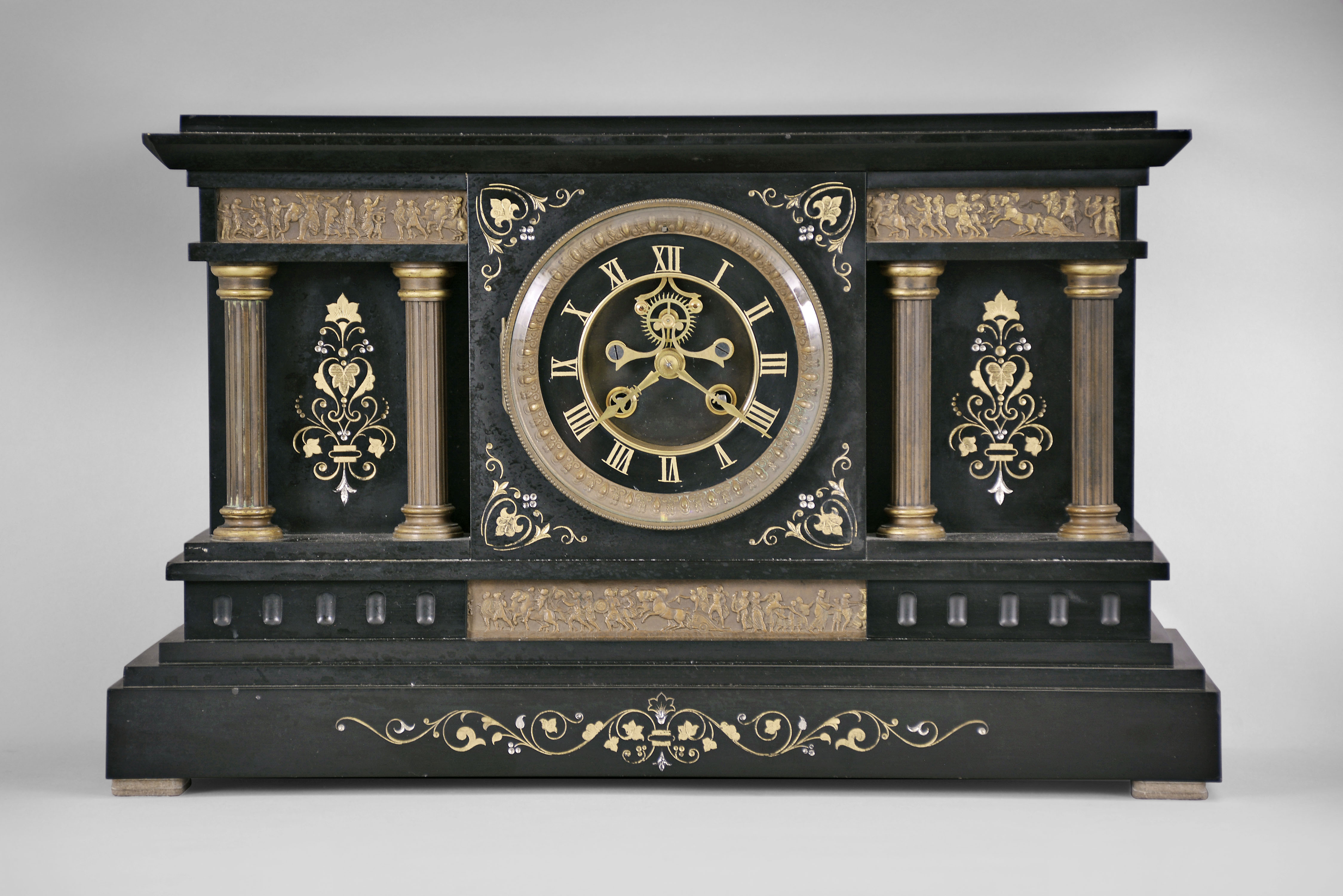 France, mantel clock, 8 day, time and strike, spring driven pendule de Paris movement with visible Brocot escapement in a black slate case with incised floral designs, gilt metal columns, and three classically inspired low-relief friezes depicting a crowd, chariot, soldiers, and an elephant, black slate dial with incised gold Roman numerals and gilt hands. — Antique Clocks Price Guide