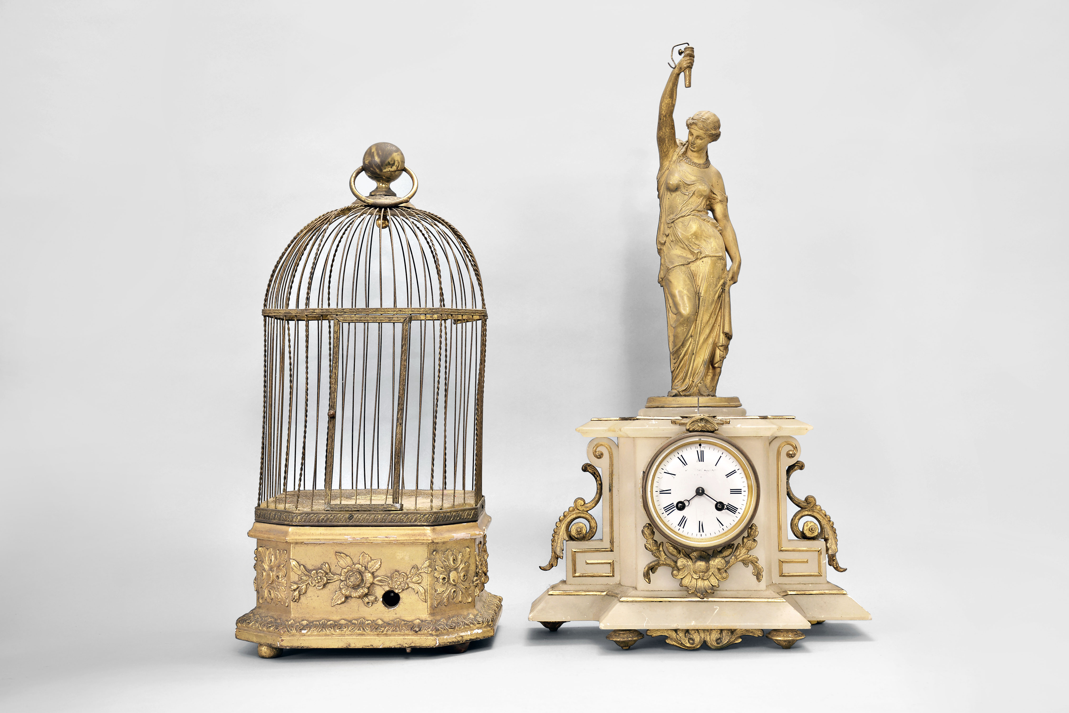 Clocks- 2 (Two): (1) France, figural mantel clock with conical pendulum, 8 day, time and strike, spring driven movement in a white stone case with applied gilt ornaments surmounted by gilt female figure holding pendulum aloft, white enamel dial with Roman numerals, and blued steel Breguet hands, c1880; (2) French, large singing bird cage, spring driven movement in a gilt wood and gesso base, brass cage, controls and winding from the base, c1880. — Antique Clocks Price Guide