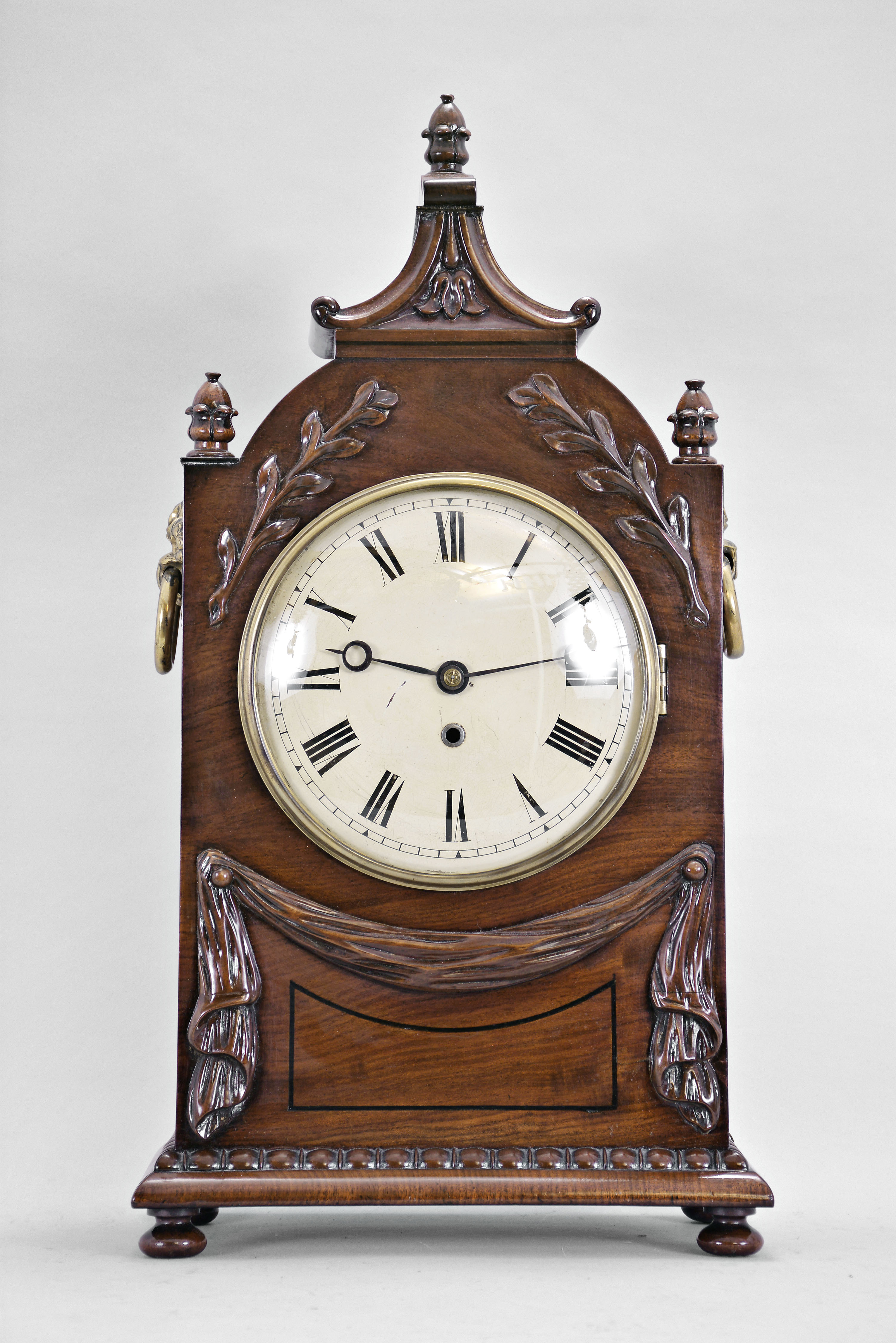 England, a 19th century mahogany bracket clock with passing strike, the case on turned wooden feet, the base with quarter round, and bead and reel moldings below a carved festoon, the upper part of the case with two carved laurel fronds, turned, carved finials, and pagoda top with carved, partial C scrolls and bellflower, the sides with pierced brass, lancet arch frets, and stamped, ring handles with floral mounts, roman numeral white painted dial with blued steel Breguet style hands, 8 day fusee timepiece with shouldered plates, recoil escapement, and passing strike at the hour, brass pendulum with brass clad bob — Antique Clocks Price Guide