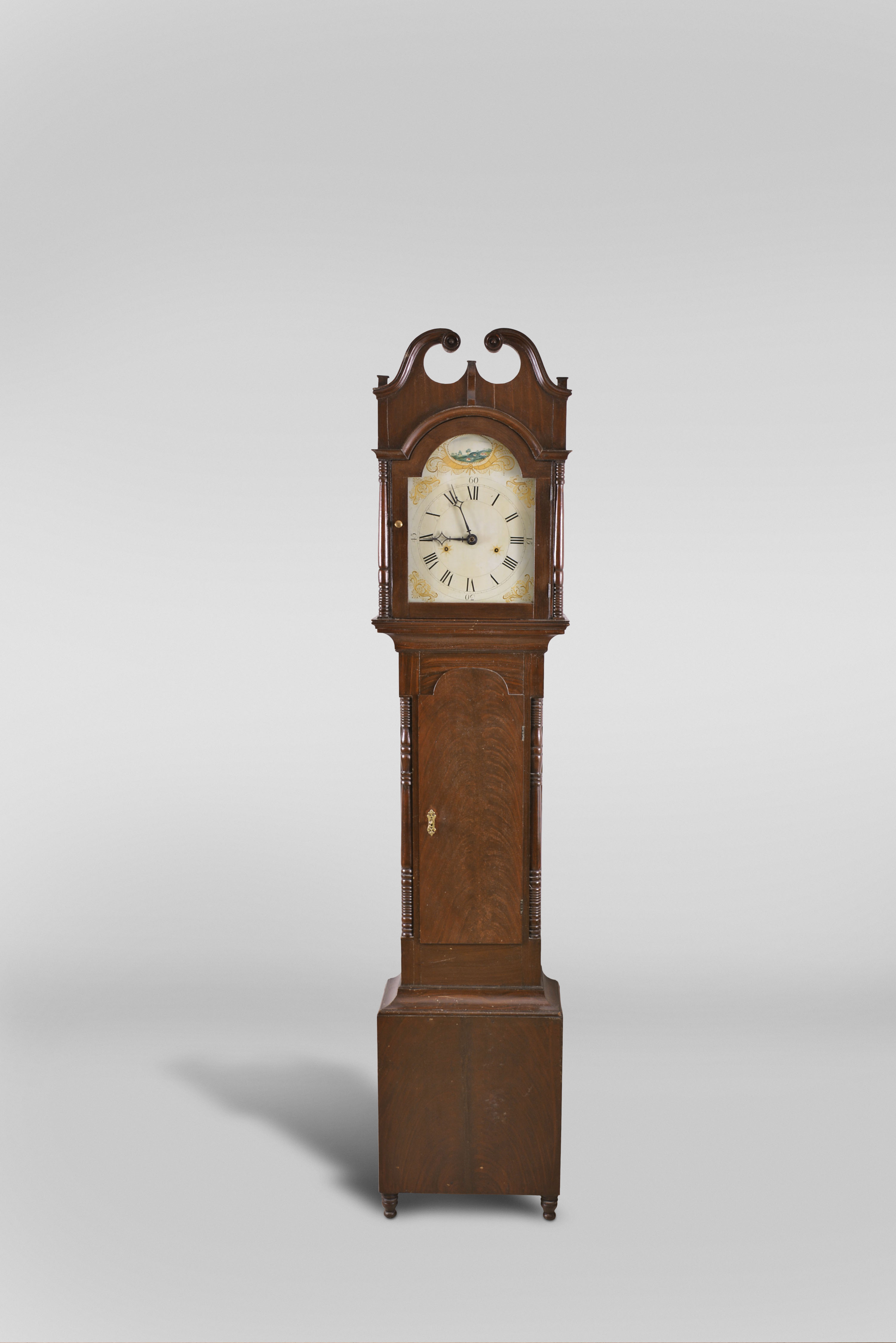 Silas Hoadley, Plymouth, Conn., tall clock, 8 day, time and strike, pull wind weight driven wooden works movement in a grain painted pine case with broken arch top, turned full columns flanking hood and trunk door resting on small turned feet. The painted wooden dial has a painted scene in the arch. — Antique Clocks Price Guide
