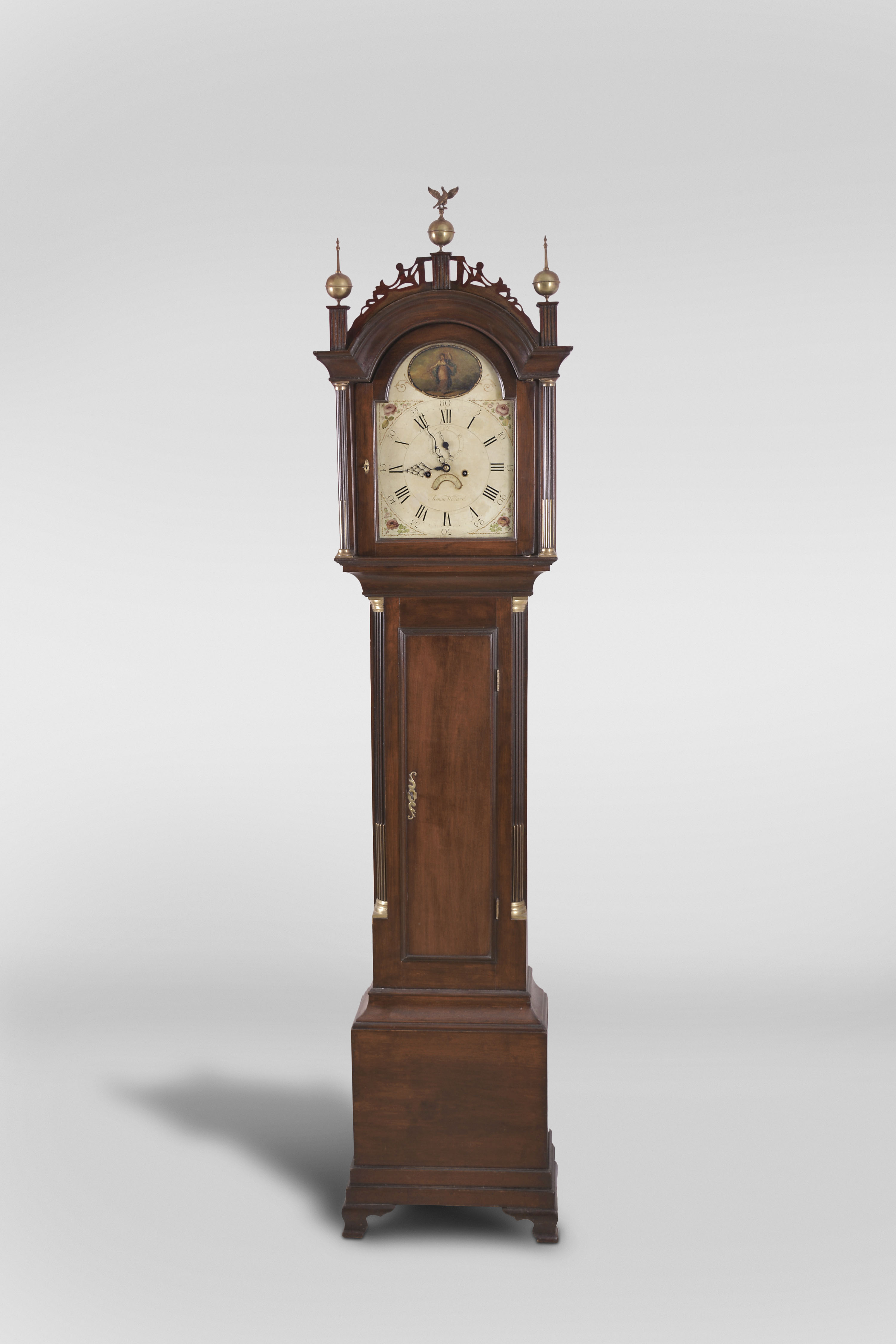 Simon Willard, Roxbury, Mass., tall clock, 8 day, time and strike, weight driven movement in typical Roxbury style mahogany case with three brass finials including center eagle finial, fretted crest, stop fluted full columns with brass capitals flanking hood and trunk doors resting on ogee bracket base. The painted metal 12-in dial by Wilson of Birmingham as found on many Willard clocks of this period is signed "Simon Willard" in script has seconds bit, calendar and scene of woman dancing in arch. — Antique Clocks Price Guide