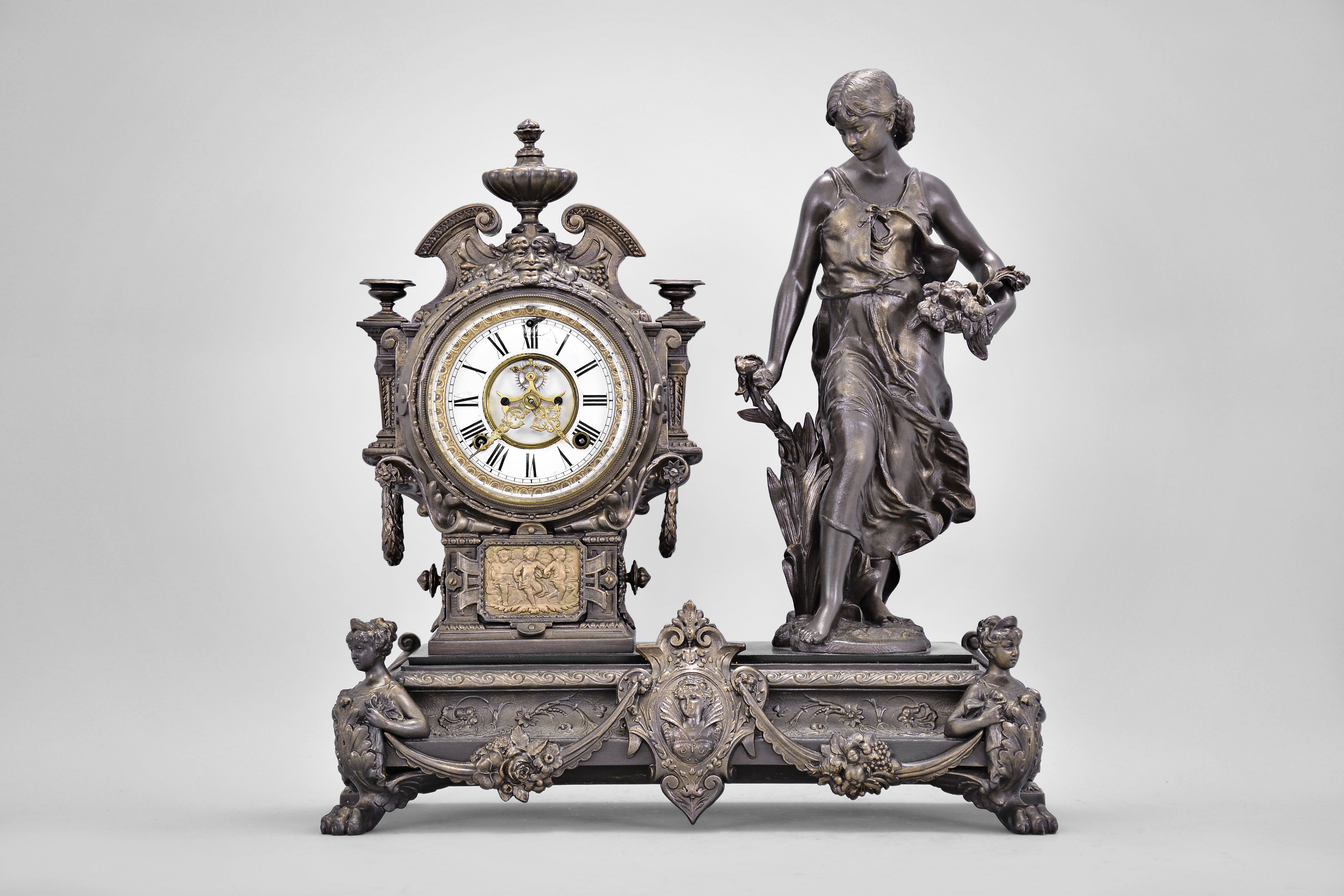 Ansonia Clock Co., New York, "Summer" figural mantel clock, 8 day, time and strike, spring driven movement with visible Brocot escapement within an ornate cast metal case, the base with paw feet and swags supporting a female figure carrying a bundle of flowers, the clock with urn top, white enamel dial, and gold Louis XV style hands. — Antique Clocks Price Guide