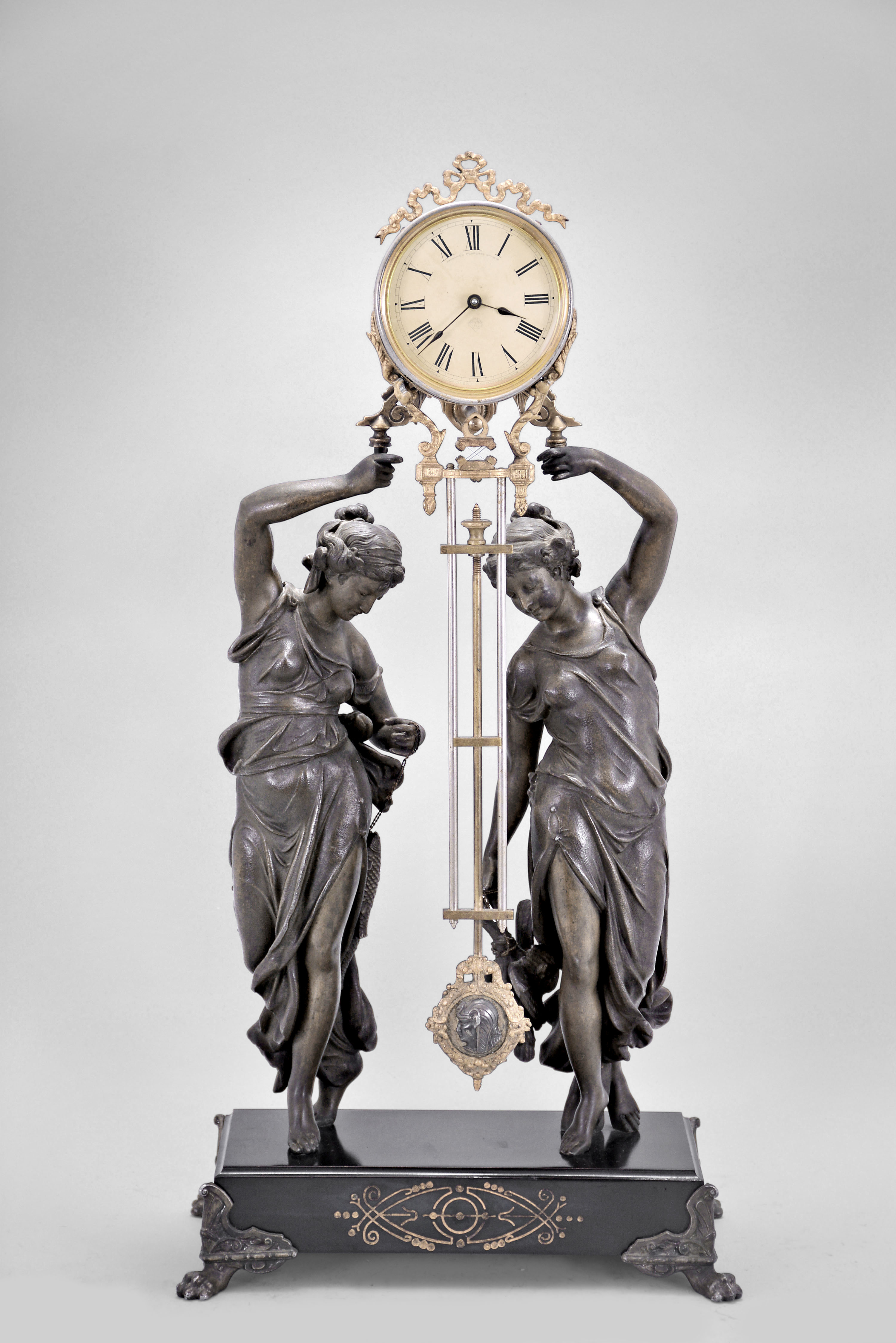 Ansonia Clock Co., New York, "Double Figure Swing" mystery clock, 8 day, time only, spring driven movement in a nickel plated cylindrical case with cast ribbon ornament, held aloft by a pair of spelter figures standing on a black enamel base with paw feet. — Antique Clocks Price Guide