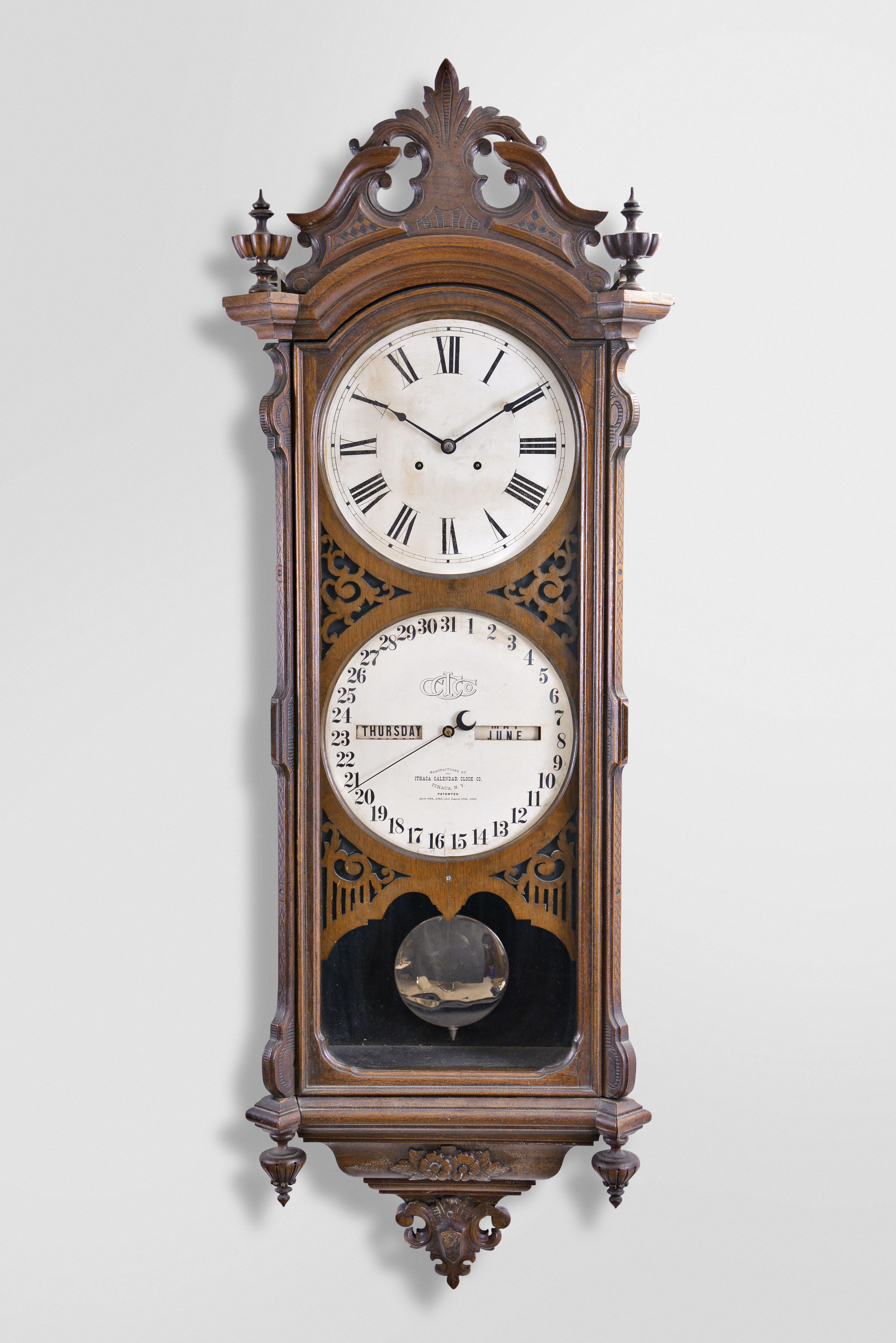 Ithaca Calendar Clock Co., Ithaca, New York, "No. 0 Bank", wall clock, 8 day weight driven movement with perpetual calendar, in a walnut case with a carved pediment top, carved base, ornately carved dial surround and upper and lower finials with papered metal dials — Antique Clocks Price Guide
