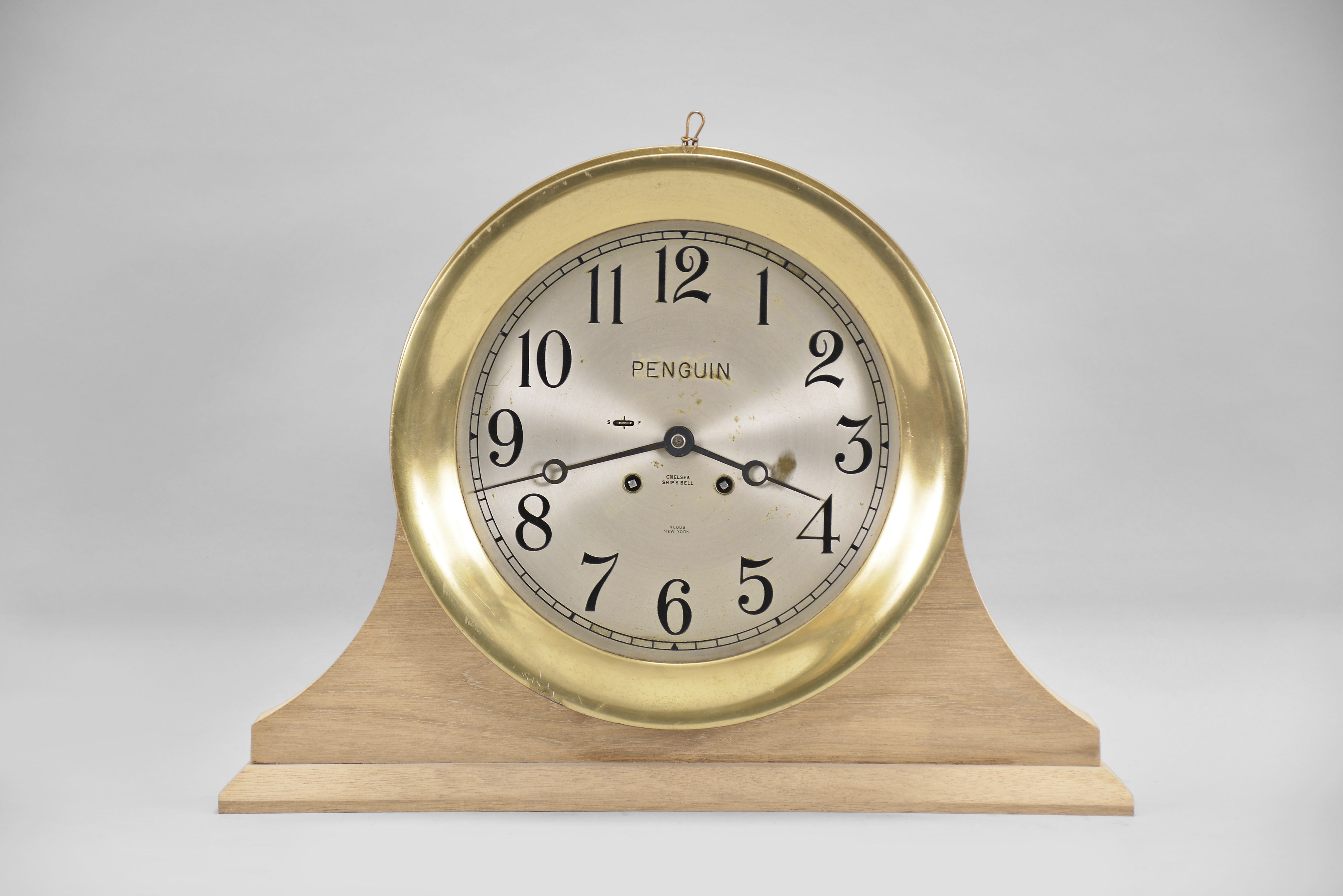 Chelsea Clock Co., Boston, Mass., "Ship's Strike" wall clock, retailed by Negus, New York for the ship "PENGUIN", 8 day, time and ship's bell strike, spring driven movement in a round brass case with 8 inch silvered metal dial accompanied by bench made wooden stand, serial #605321 — Antique Clocks Price Guide