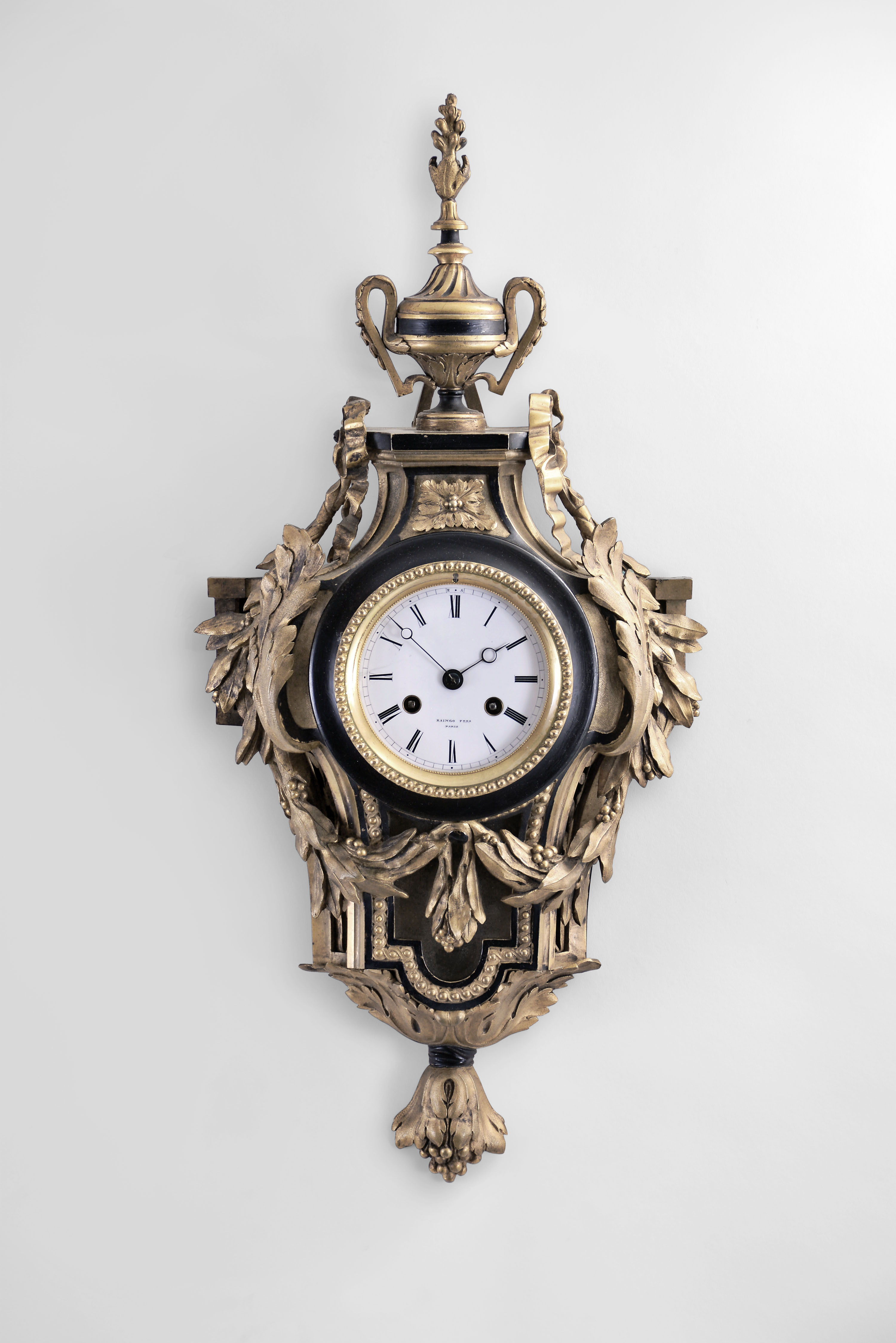Raingo Freres, Paris, cartel clock, the gilt bronze case with classical ornament, the top with urn finial above flanking bow knots at the sides, issuing a pair of laurel garlands, the festoons pinned together at the center below the dial and terminating before the guilloche molded pendulum aperture, the laurel partially obscured by a pair of upturned acanthus leaves, and the base with bound acanthus leaf and pendant husk drop, the gilt, beaded bezel with beveled glass, Roman numeral white enamel dial, blued steel Breguet style hands, 8 day time and strike pendule de Paris movement sounding the hour and half hour on a bell, dial and movement signed — Antique Clocks Price Guide