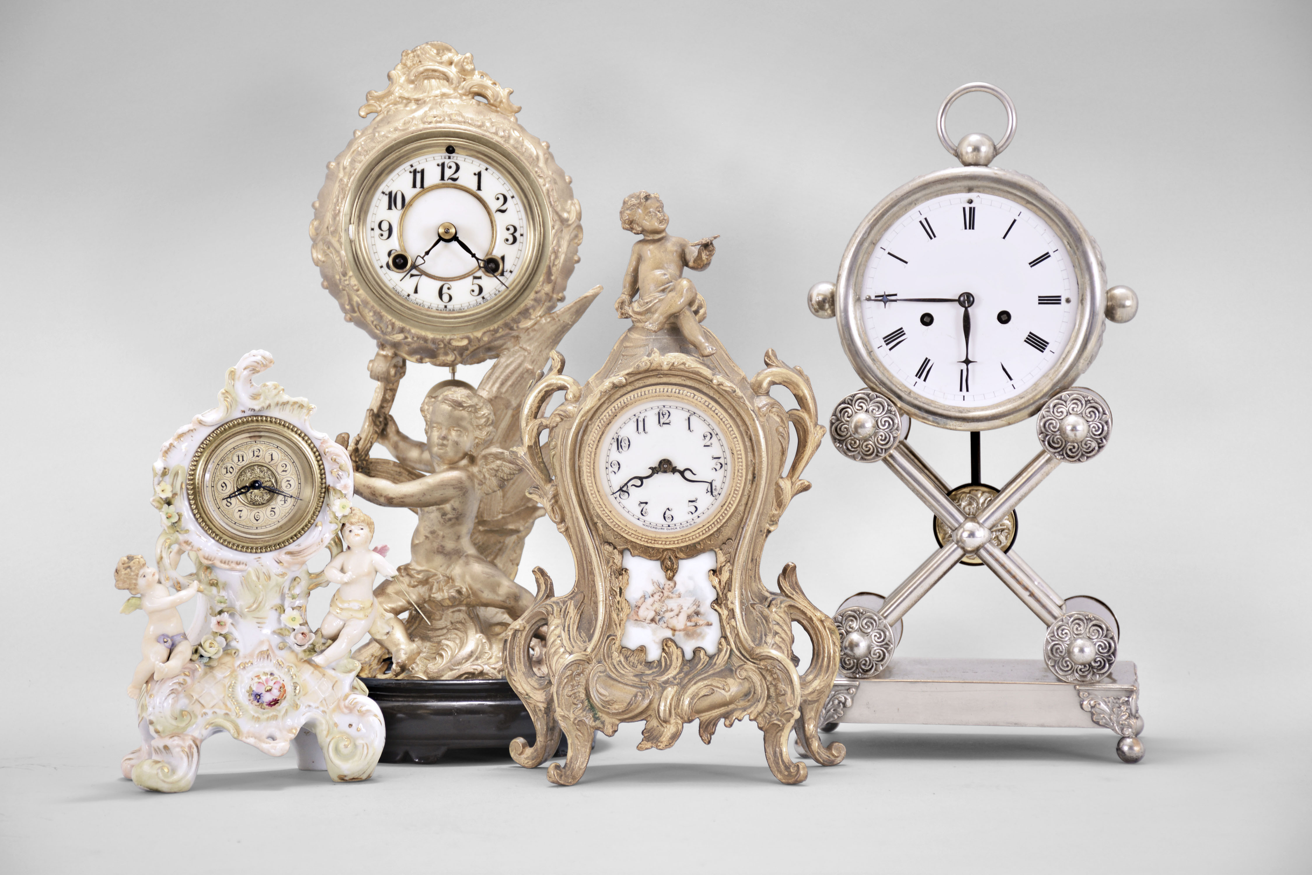 Clocks- 4 (Four): (1) Waterbury Clock Co., Waterbury, Conn., mantel clock, 8 day, time and strike, spring driven movement in a cast metal case featuring a putto holding the clock aloft, cream porcelain dial, 1915; (2) Austria, mantel clock, 2 day, time and strike, spring driven movement in a silver plate-over-copper case with white enamel dial, c1840; (3) Waterbury Clock Co., Waterbury, Conn., novelty clock, 30 hour, time only movement with thread suspension in a cast metal case with painted ceramic panel and topped by putto figure, c1905; (4) Germany, novelty clock, 30 hour, time only, spring driven movement in a porcelain case with putti figures, c1920. — Antique Clocks Price Guide