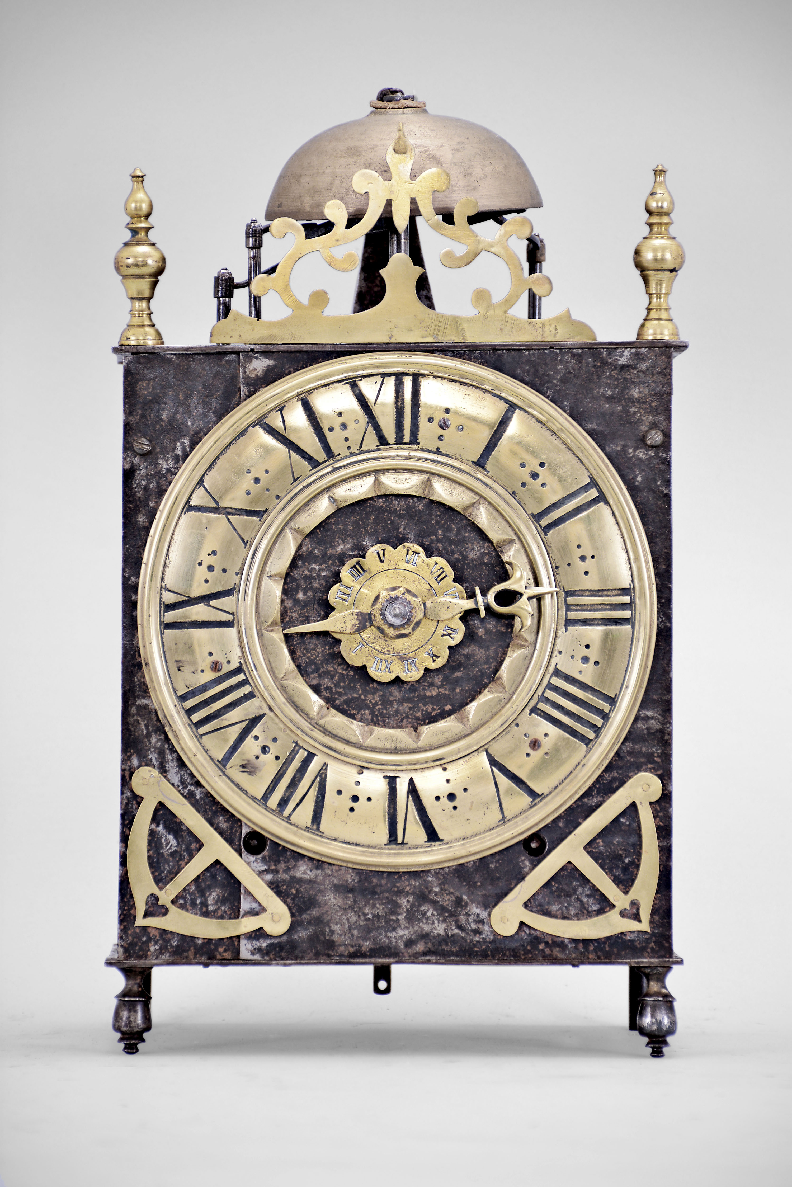 France, Comtoise lantern clock, 8 day, time and strike with alarm, weight driven movement with verge escapement, thread suspension with rear-hanging pendulum in a posted iron frame case, heavy brass chapter ring with Roman numerals, single brass hand, alarm ring with Roman numerals, applied brass spandrels with hearts, all resting on turned iron feet in the front, and topped with a thick pierced brass fronton flanked by two turned brass finials. — Antique Clocks Price Guide