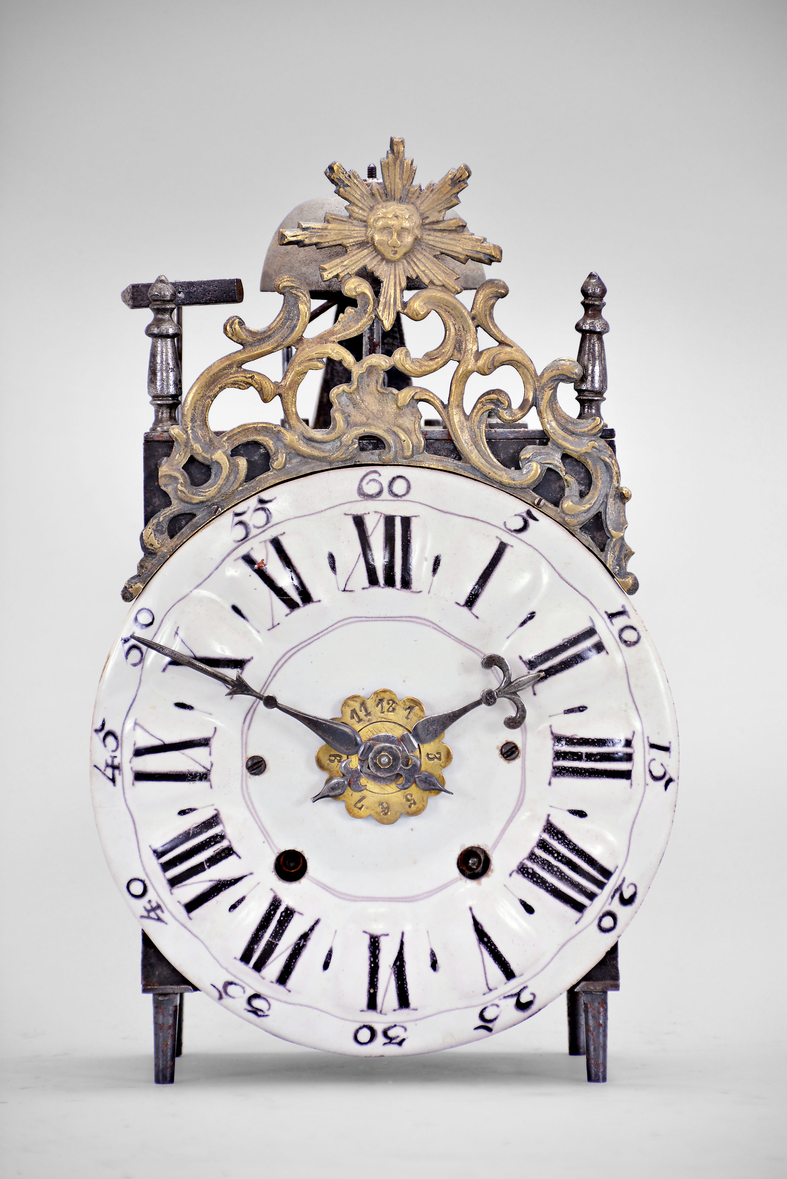 France, Comtoise lantern clock, 8 day, time and strike with alarm, weight driven movement with verge escapement, thread suspension with rear-hanging pendulum in an iron post and plate frame, early faience dial with raised portions under each Roman numeral, outer ring with Arabic numerals every five minutes, two steel hands, brass alarm ring with Arabic numerals, topped by a cast brass fronton with image of the sun / Apollo flanked by two turned iron finials, all standing on tapered iron feet. — Antique Clocks Price Guide