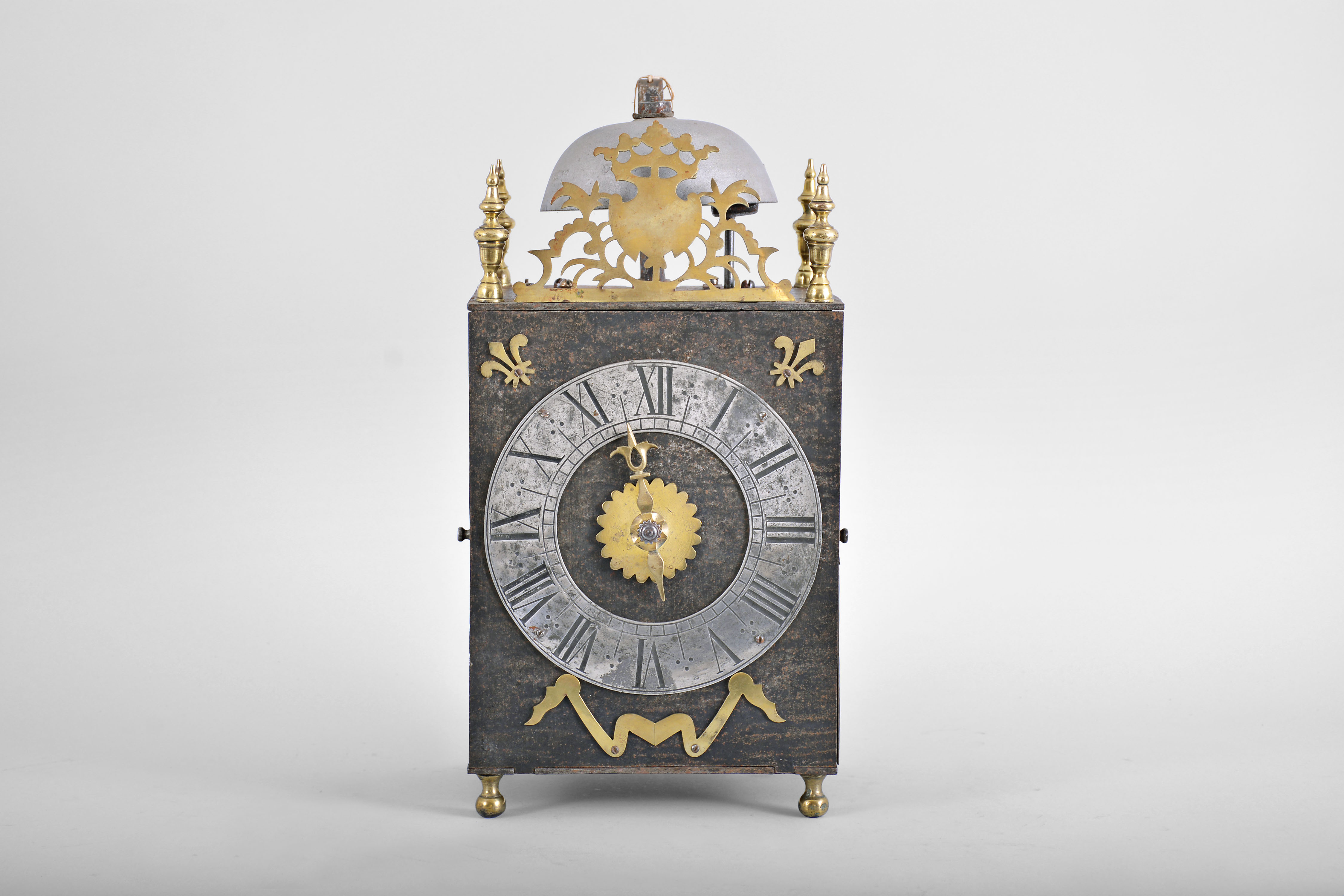 France, Comtoise lantern clock, 8 day, time and strike, weight driven movement with verge escapement, thread suspension with rear-hanging pendulum in a iron post and plate frame, pewter chapter ring with engraved Roman numerals, single brass hand, applied brass fleur de lis spandrels and pivoted winding arbor shutters, all resting on four turned brass feet, and topped by flat pierced brass fronton flanked by four turned brass finials. — Antique Clocks Price Guide