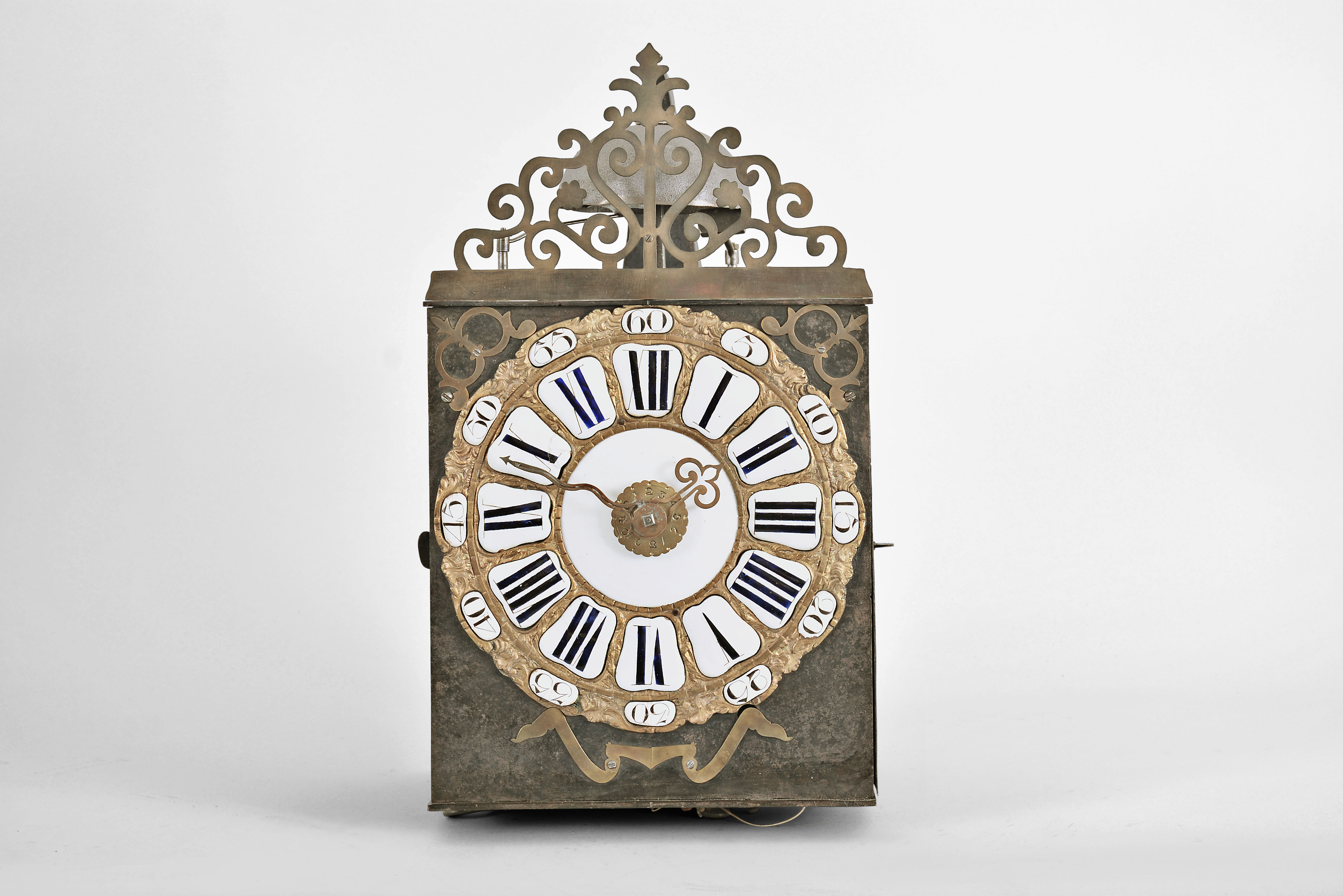 France, month-running Comtoise clock, 30 day, time and strike with alarm, weight driven movement, verge escapement with thread suspension and rear-hanging pendulum in an iron post and plate frame, cast brass dial with inset white enamel cartouche numerals with blue Roman numerals, outer ring with Arabic numerals every five minutes, brass alarm ring with Arabic numerals, two brass hands, applied brass spandrels and pivoted winding arbor shutters, all topped by a pierced brass fronton. — Antique Clocks Price Guide