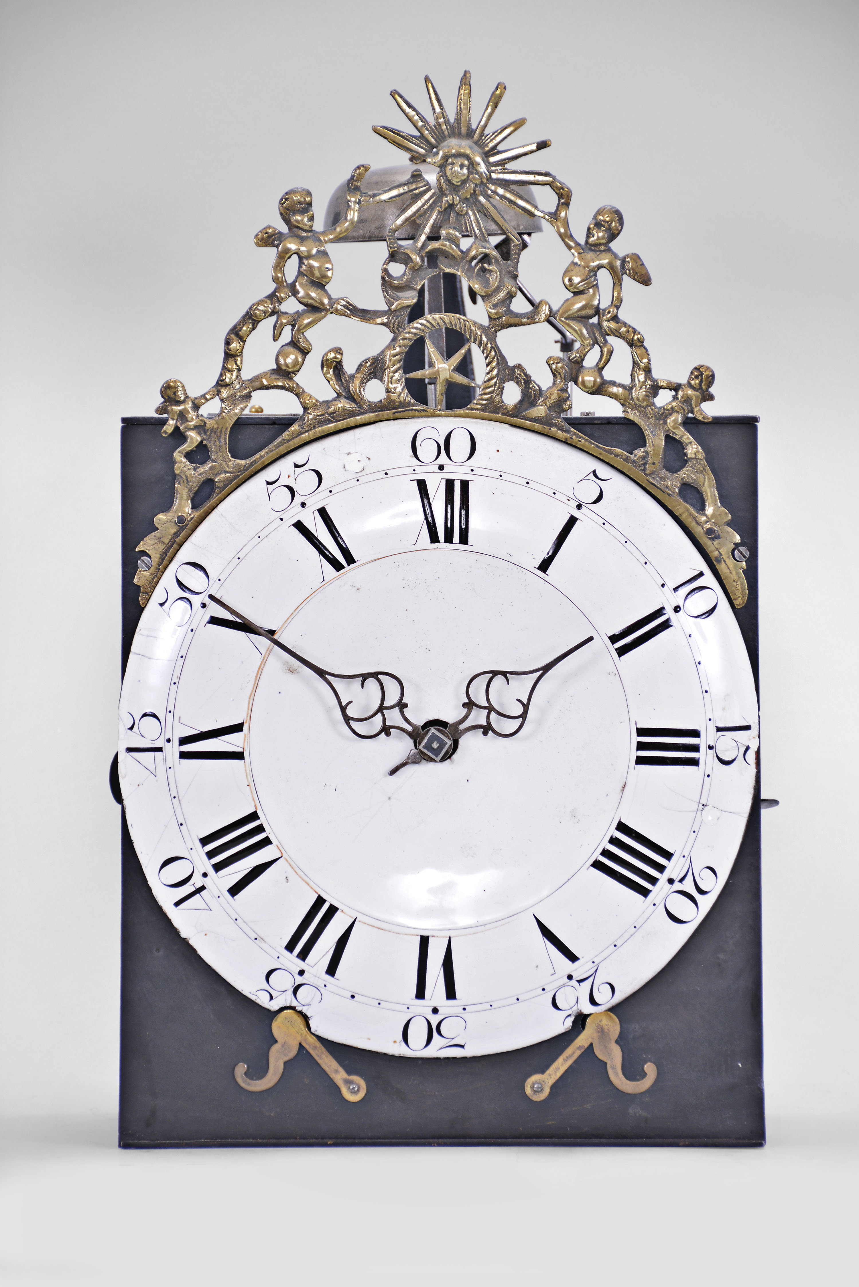 France, month-running Comtoise clock, 30 day, time and strike, weight driven movement, verge escapement with thread suspension and rear-hanging pendulum in an iron post and plate frame, white enamel dial with concave center and convex chapter, Roman numerals for the hours and Arabic numerals every five minutes, two pierced iron hands, pivoted brass winding arbor shutters below the dial, all topped by a cast fronton featuring the sun flanked by putti and displaying the star symbol used during the French Restoration Period. — Antique Clocks Price Guide