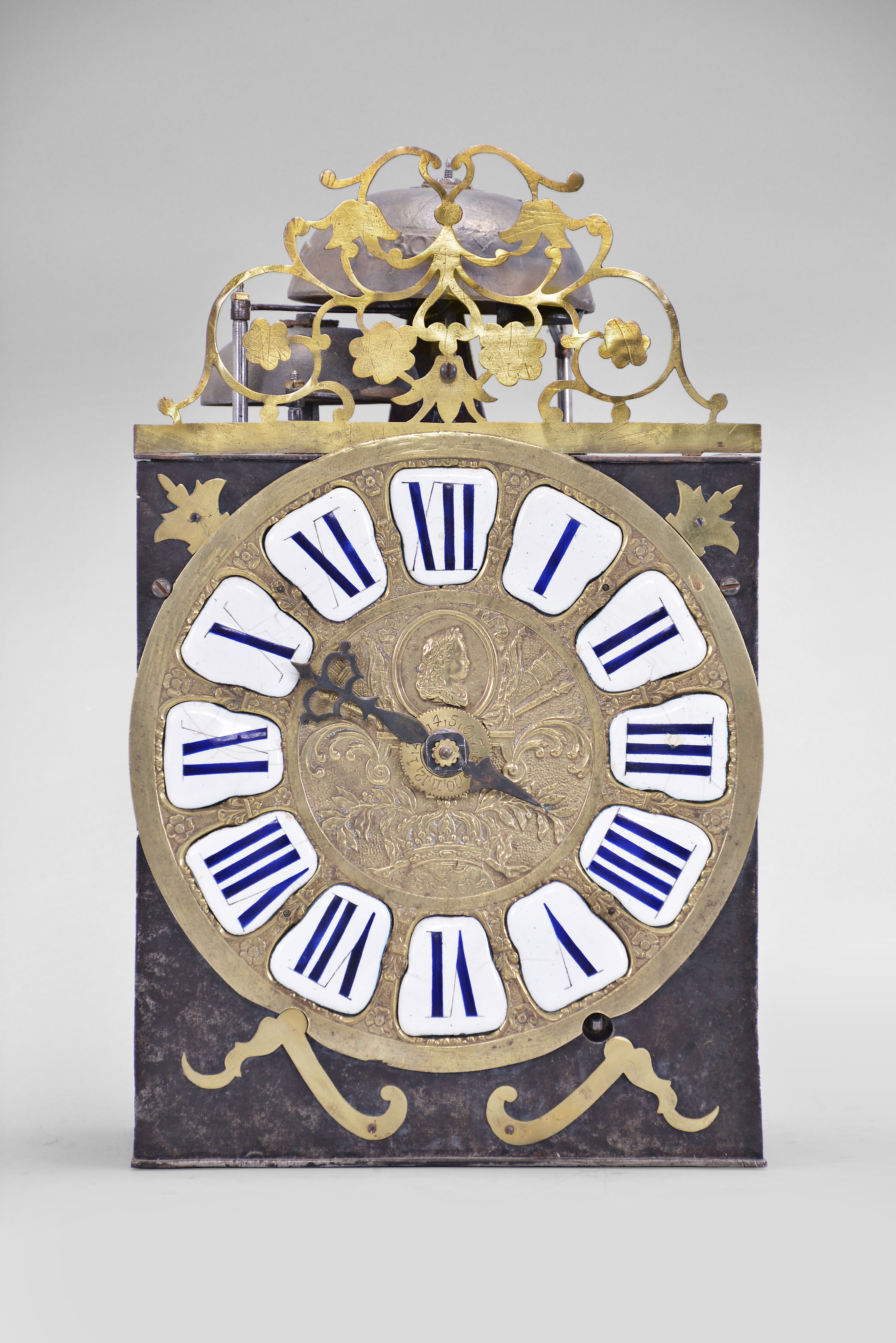France, Comtoise clock, 8 day, time and strike with alarm, weight driven movement with rare Maillet (or Mayet) escapement and unusual double vertical rack strike in a posted iron frame, cast brass dial with profile of young Louis XV with inset white enamel cartouches with blue Roman numerals, alarm ring with Arabic numerals, single pierced iron hand, brass spandrels above and pivoted winding arbor shutters below, all topped with a pierced brass fronton with floral motif. — Antique Clocks Price Guide