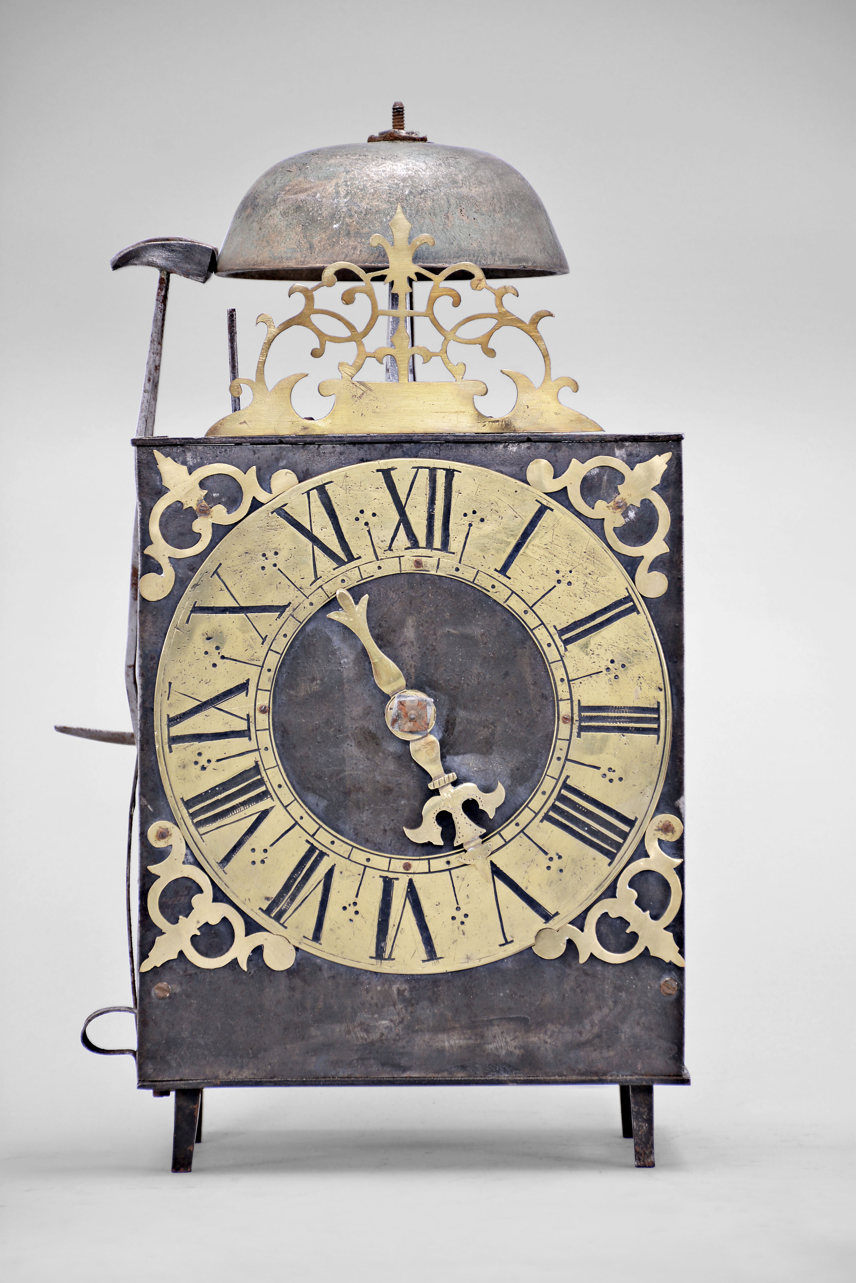 France, Comtoise lantern clock, 30 hour, time and strike, weight driven movement with rare Maillet or Mayet escapement and atypical vertical rack strike with side mounted hammers, iron post and plate frame construction, brass dial with engraved Roman numerals, single brass hand, brass spandrels above and pivoted winding arbor shutters below, all topped with a pierced brass fronton with fleur de lis at the apex. — Antique Clocks Price Guide