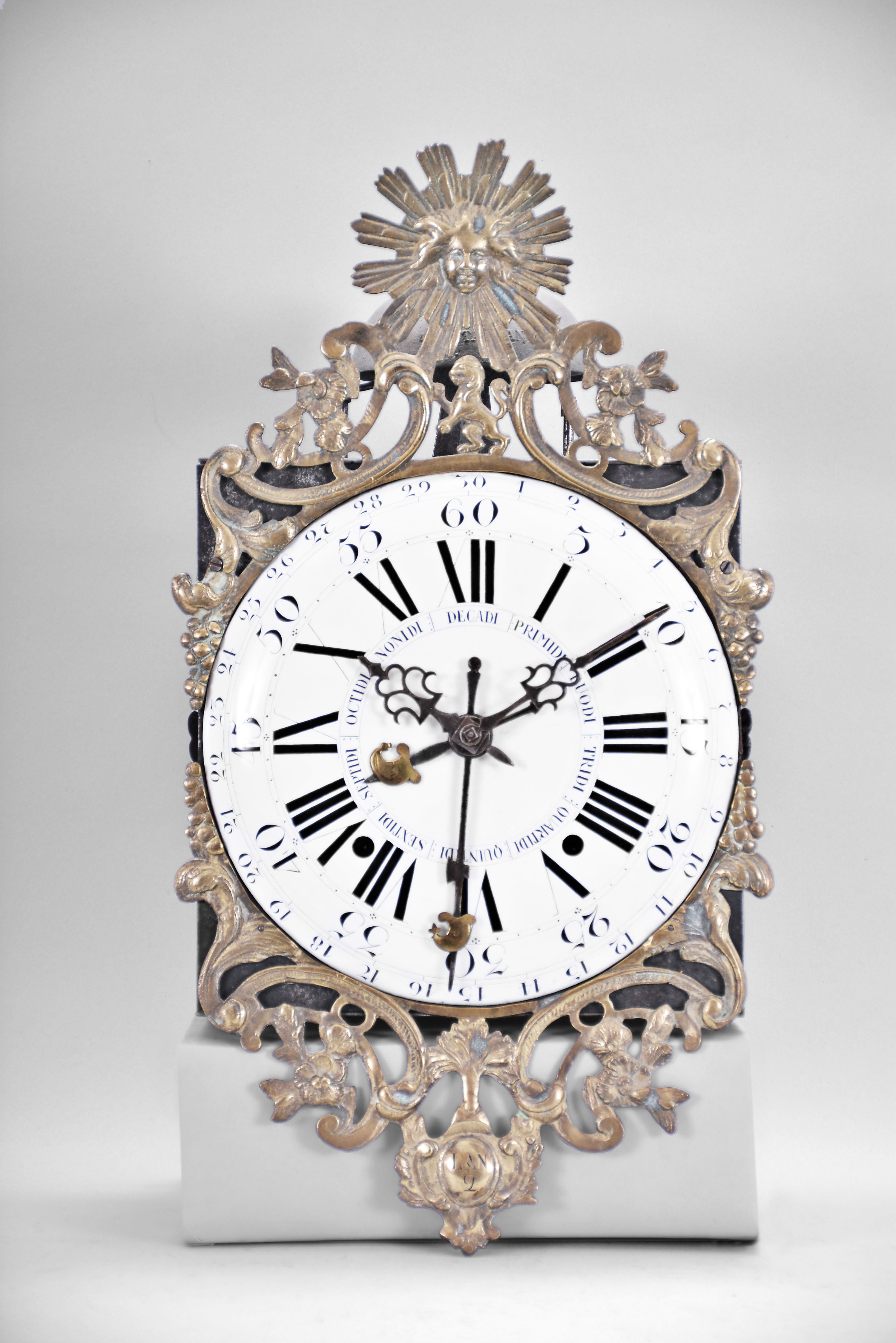 France, Revolutionary Era bronze-front Comtoise clock, 8 day, time and strike, day of the month and day of the week according to the French Republican Calendar, weight driven movement with verge escapement, thread suspension with rear-hanging pendulum in an iron post and plate frame, early enamel dial with concave center and convex chapter, Roman numerals for the hours, Arabic numerals for the minutes and days of the month, four iron hands, date hands embellished with Phrygian caps, cast bronze fronton with sun and lion motifs, lower cast fronton with medallion engraved with the date. — Antique Clocks Price Guide
