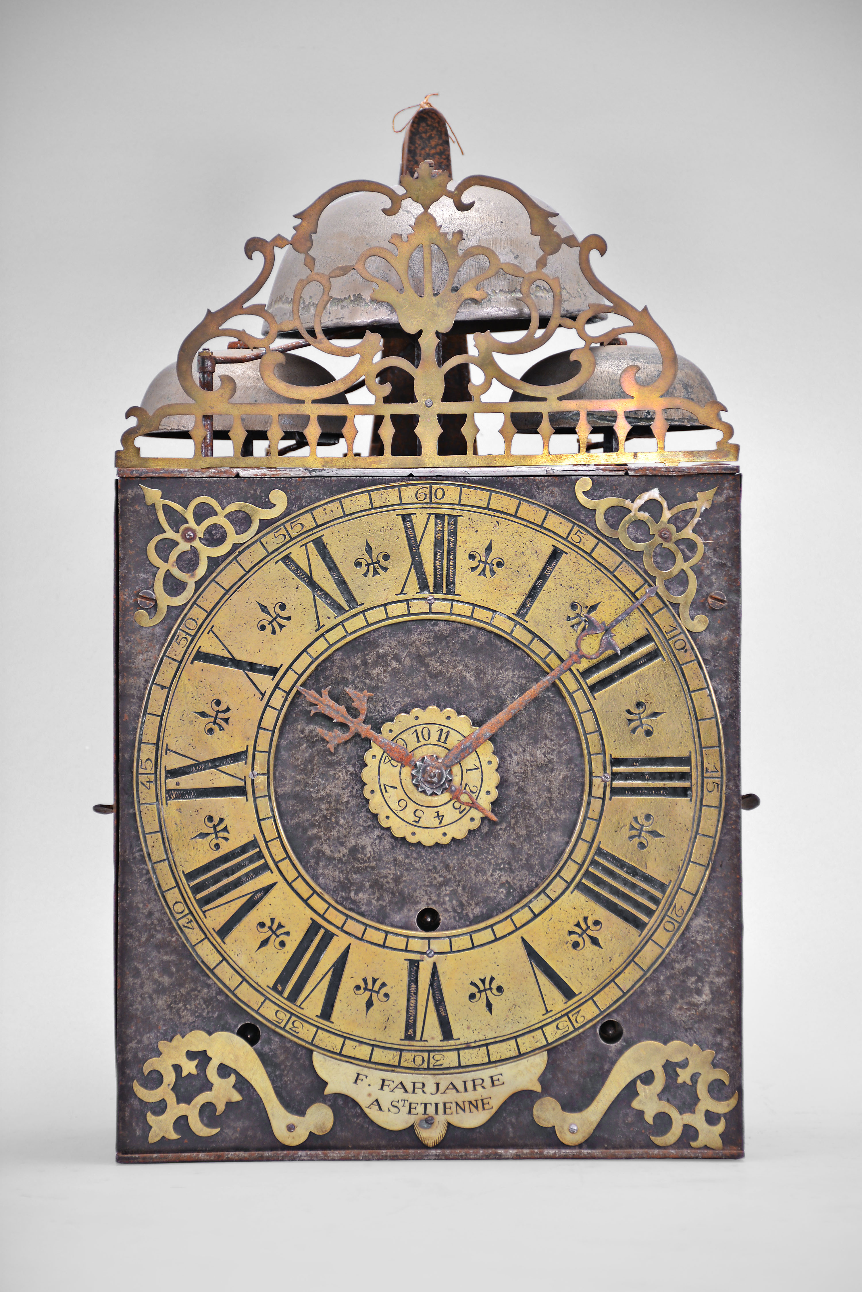 F. Farjaire, Saint-atienne, France, three train Comtoise clock, 8 day, time and quarter strike with alarm, three bells, weight driven movement with verge escapement, thread suspension with rear-hanging pendulum in an iron post and plate frame, brass chapter with Roman numerals, Arabic numerals at 5 minute intervals on the outer rim, brass alarm ring with Arabic numerals, two iron hands, pivoted brass winding arbor shutters, engraved plaque with the maker's name, all topped by pierced brass fronton. — Antique Clocks Price Guide