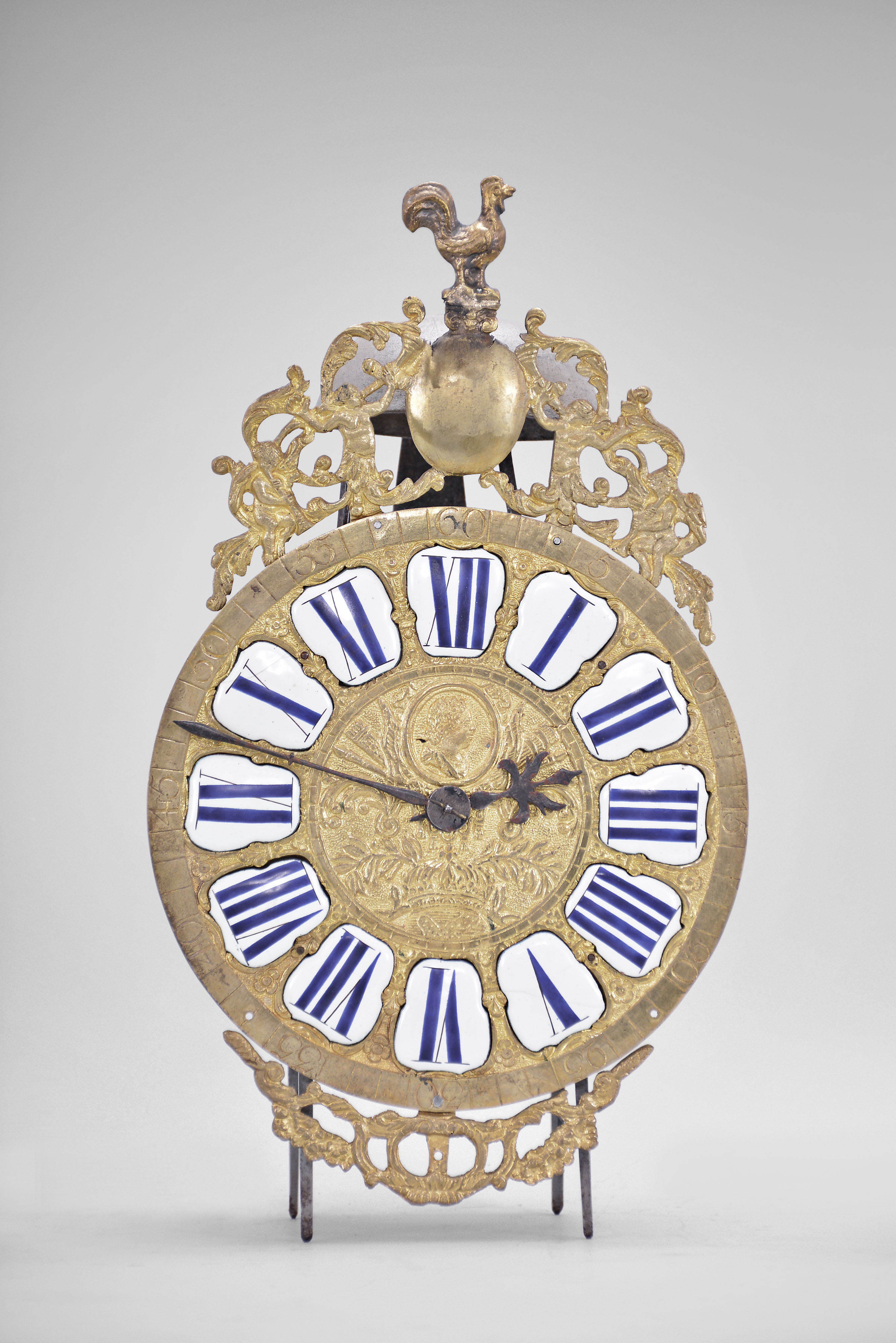 France, Comtoise lantern clock, 30 hour, time and strike, weight driven movement with rare pin wheel escapement, iron frame and feet, rear mounted thread suspension, cast brass dial with profile of Louis XV and inset with 12 white enamel cartouches with Roman numerals, outer ring with Arabic numerals every 5 minutes, cast bronze fronton with trumpeters flanking a medallion topped by the Gallic rooster, two iron hands. — Antique Clocks Price Guide