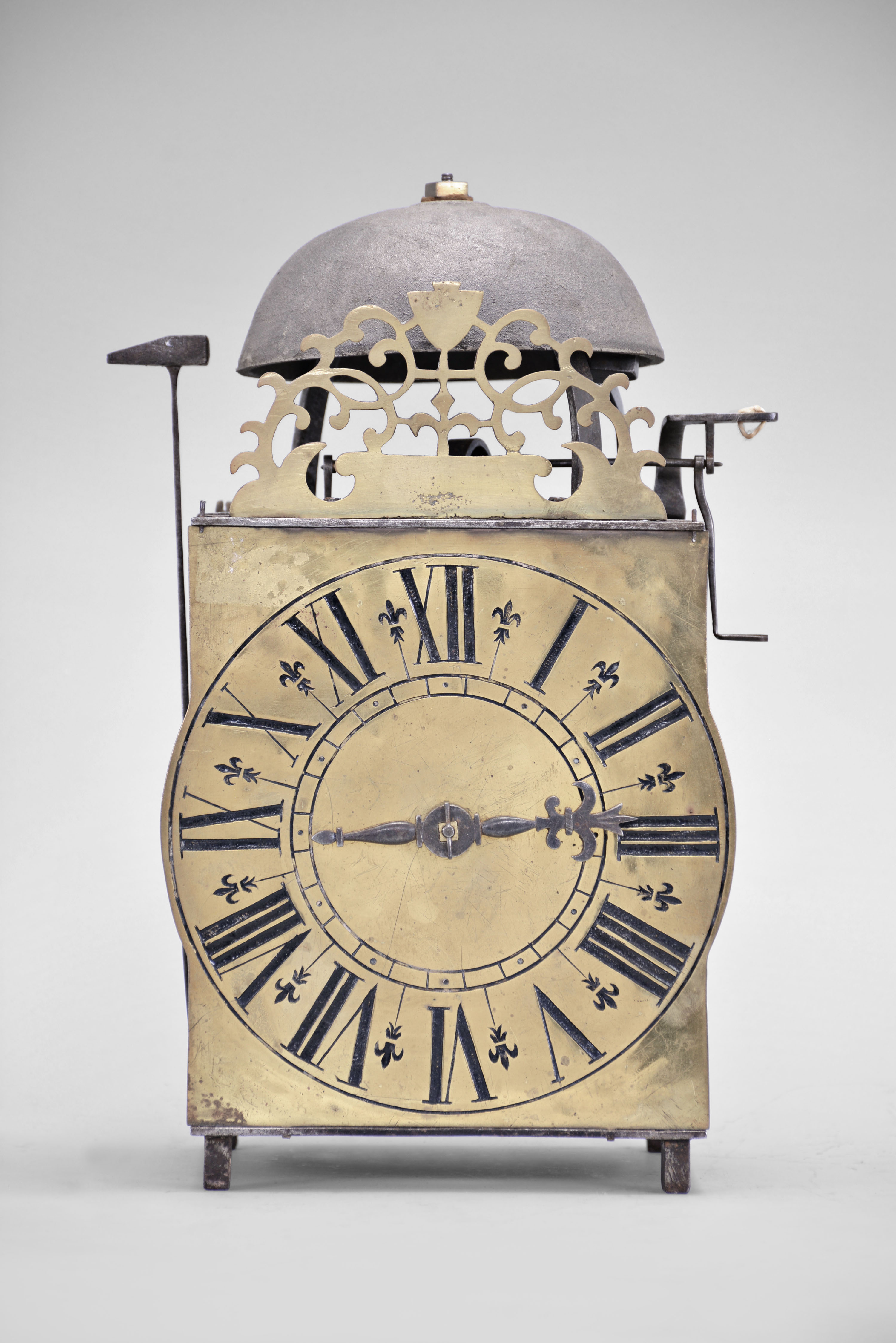 France, lantern clock, 30 hour, time and strike, weight driven movement, verge escapement with upward facing crown wheel, thread suspension with side-mounted pendulum, posted iron frame resting on short iron feet, brass front with integrated dial showing Roman numerals, single iron hand, topped by bell and pierced brass fronton. — Antique Clocks Price Guide