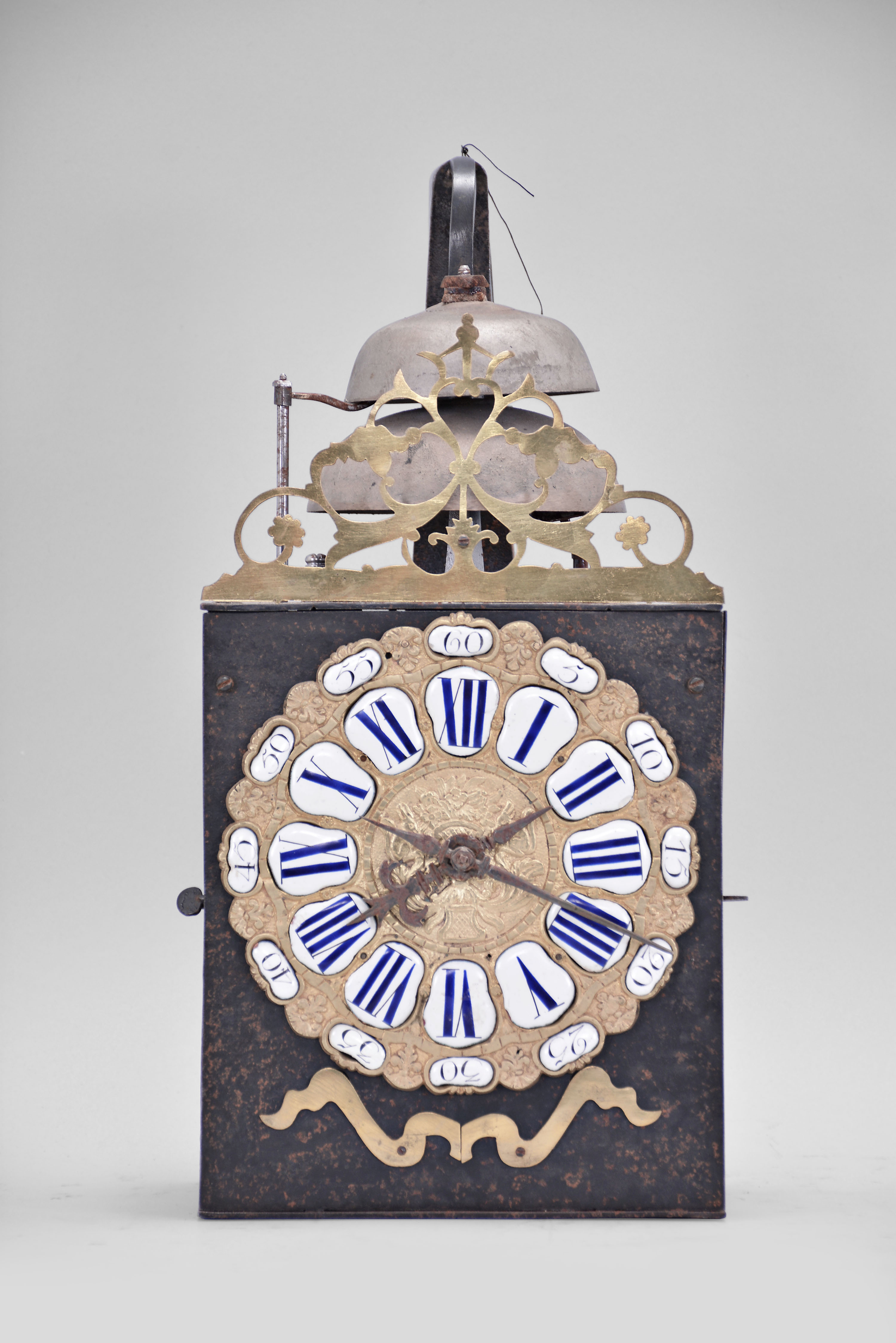 France, Comtoise clock, 8 day, time and strike with two bells, weight driven movement, verge escapement, thread suspension with rear-hanging pendulum in an iron post and plate frame, cast brass dial with two iron hands, floral motif and inset with 24 white enamel cartouche numerals, blue Roman numerals for the hours and black Arabic numerals every five minutes, flat brass winding arbor shutters below the dial, and pierced brass fronton. — Antique Clocks Price Guide