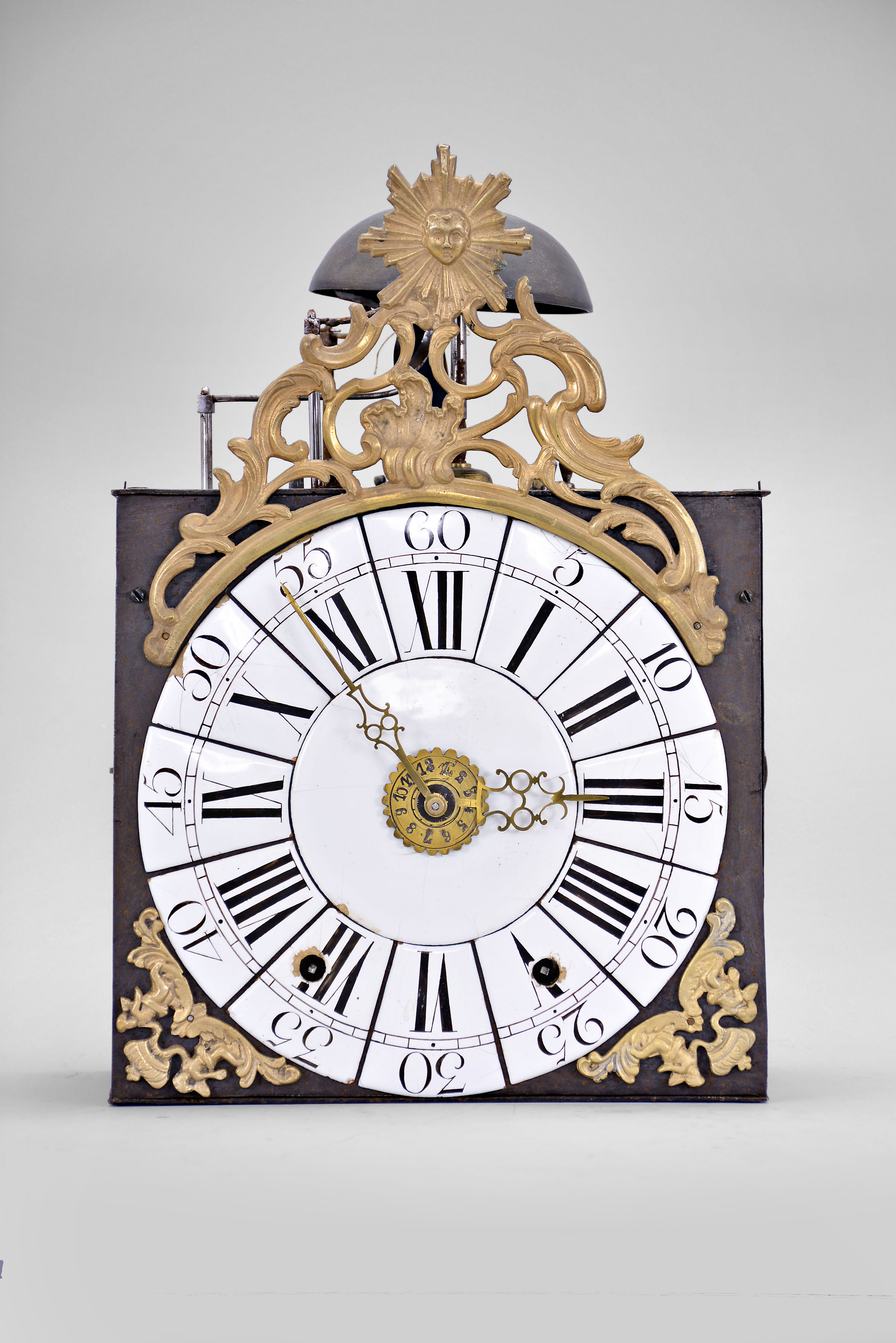 France, Louis XV Comtoise clock, 8 day, time and strike with alarm, weight driven movement with verge escapement, thread suspension with rear-hanging pendulum, iron post and plate frame, dial composed of 13 enamel panels with Roman and Arabic numerals, brass alarm ring with Arabic numerals, two pierced brass hands, cast brass spandrels below the dial with putti supporting crowns, all topped by a cast brass fronton with sun emblem within a rococo design. — Antique Clocks Price Guide