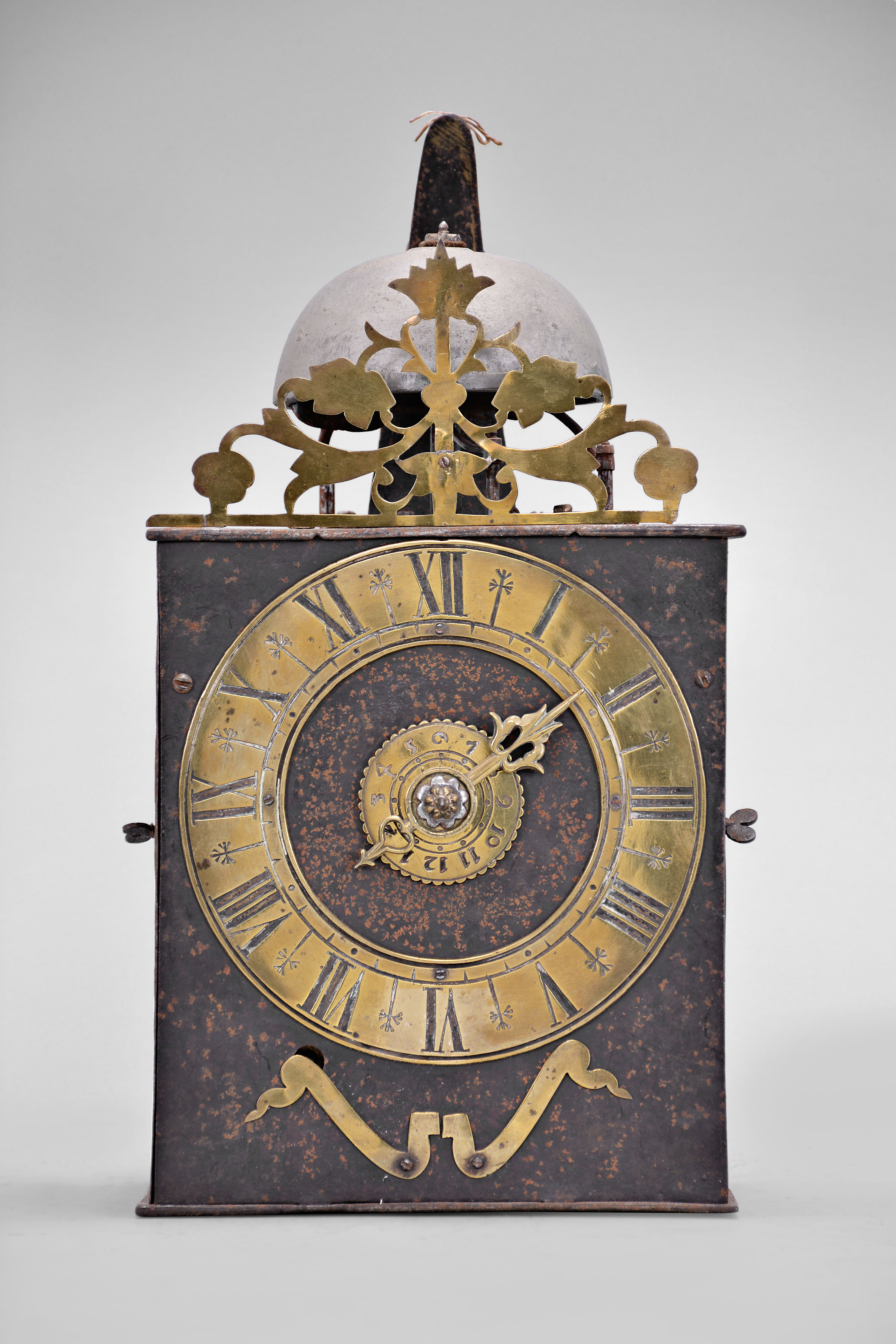 France, Comtoise clock, 8 day, time and strike with alarm, weight driven movement with verge escapement, thread suspension with rear-hanging pendulum in a iron post and plate frame, brass chapter ring with Roman numerals, brass alarm ring with Arabic numerals, pivoted winding arbor shutters, single pierced brass hand, all surmounted by a pierced brass fronton. — Antique Clocks Price Guide