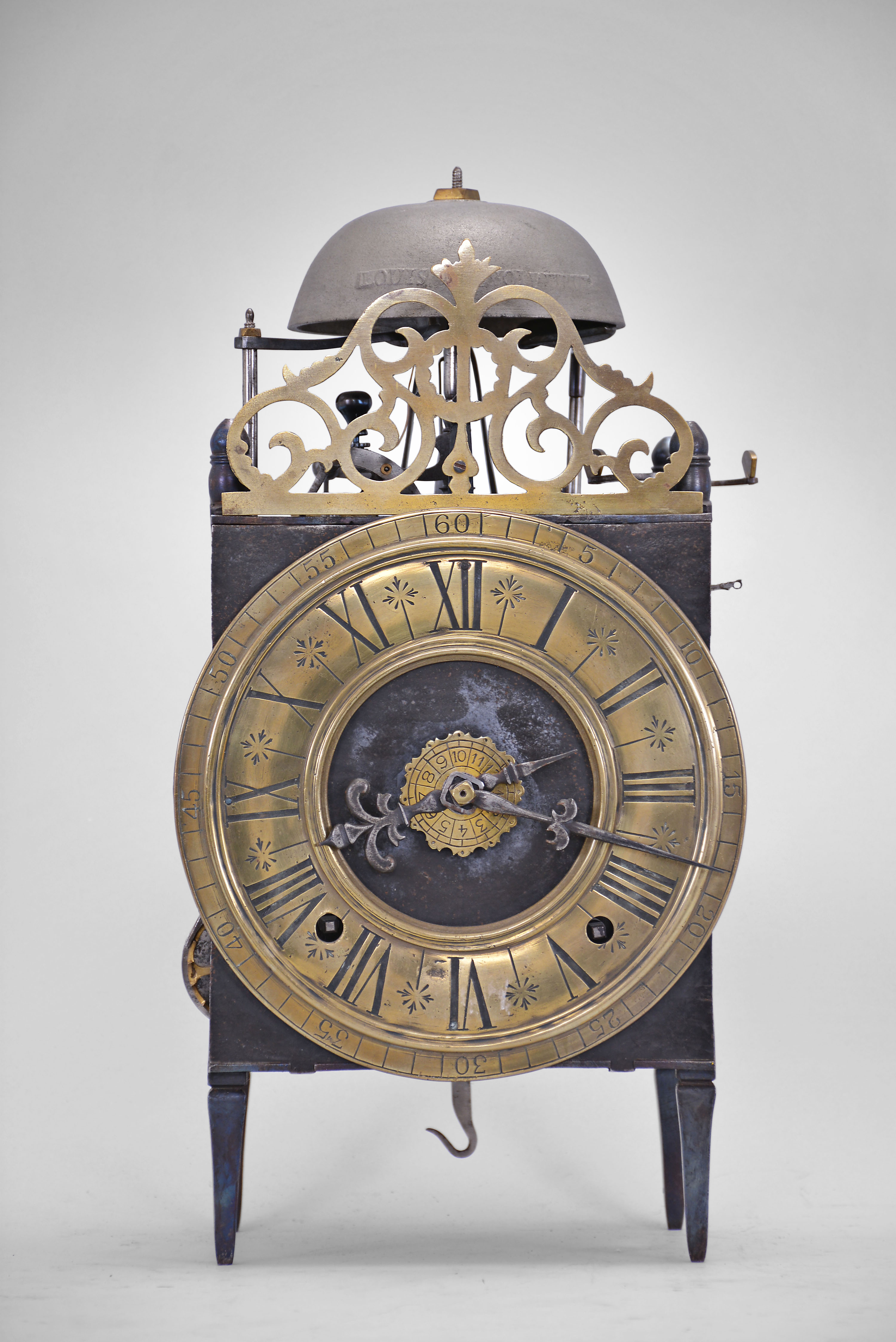 France, Comtoise lantern clock, 8 day, time and strike with alarm, weight driven movement with Chevalier de Bethune escapement, thread suspension with rear-hanging pendulum in a iron posted iron frame, brass chapter ring with engraved Roman numerals, brass alarm ring with engraved Arabic numerals, two iron hands, all surmounted by a cast brass fronton and four turned iron finials. — Antique Clocks Price Guide