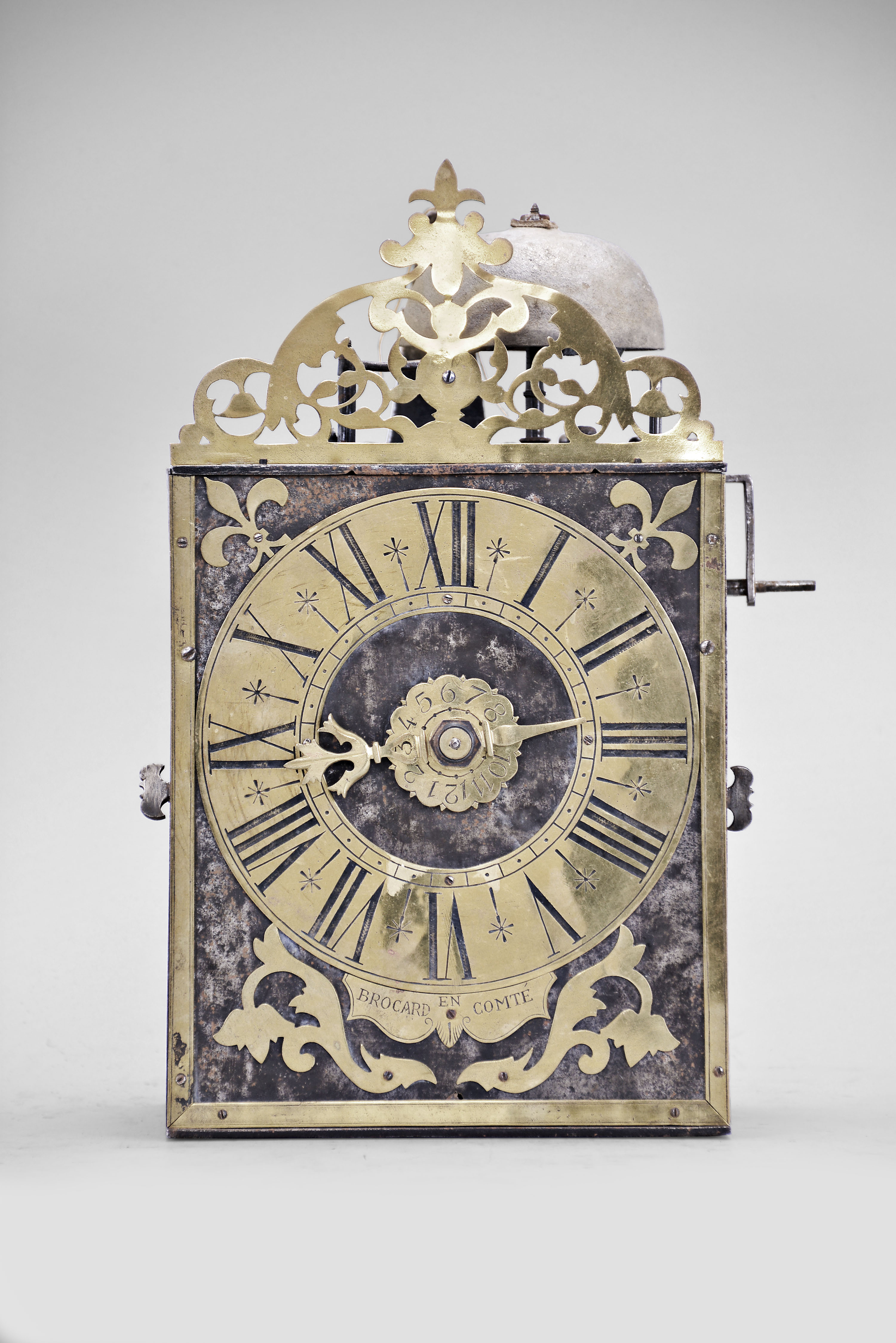 Brocard, Comte, France, Comtoise clock, 8 day, time and alarm, weight driven movement with verge escapement, thread suspension with rear-hanging pendulum in a iron post and plate frame, brass chapter ring with engraved Roman numerals, brass alarm ring with engraved Arabic numerals, flat brass fleur de lis spandrels and pivoted winding arbor shutters, engraved plaque below the dial reading "Brocard en Comte", single pierced brass hand, all topped with a pierced brass fronton. — Antique Clocks Price Guide