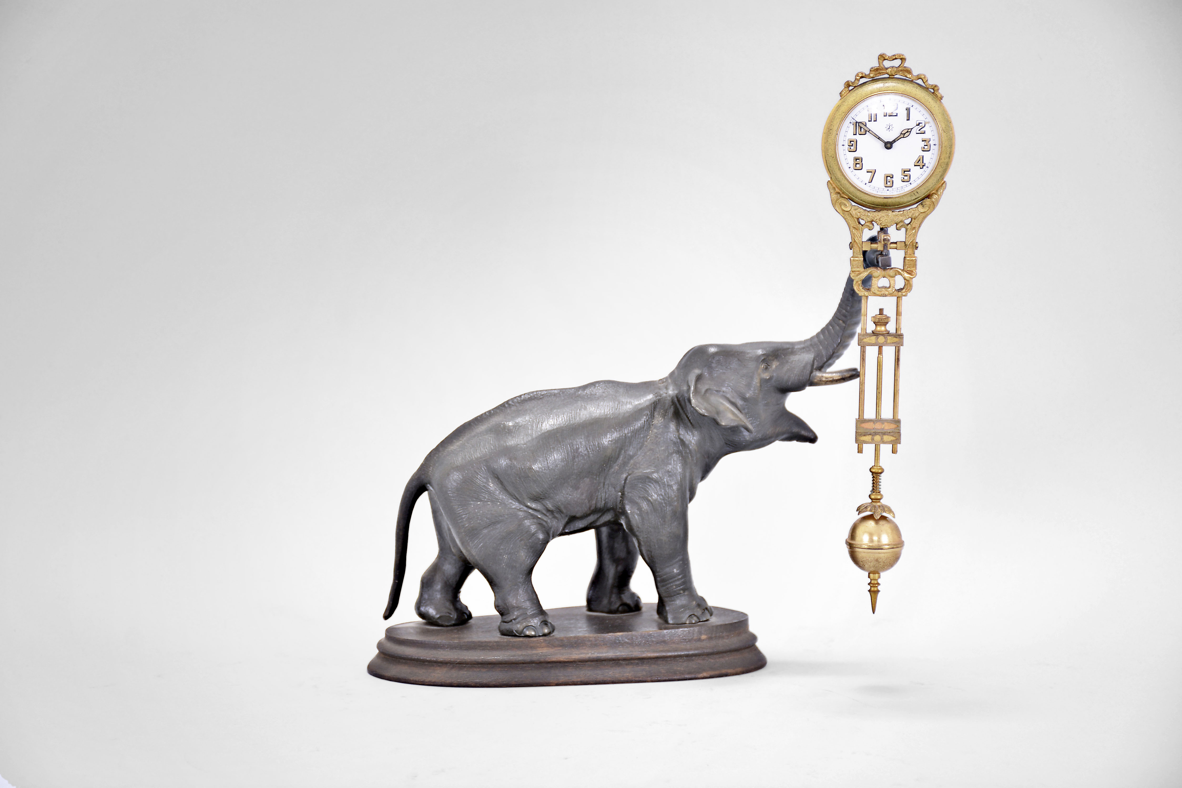 Gebruder Junghans, Schramberg, Germany, elephant model swinging clock, 8 day, time only, spring driven movement with compound pendulum held aloft by a patinated elephant figure on a molded wood base, the clock case with gilt and patinated surfaces, two-tone Arabic numerals on a white enamel dial with the Junghans star trademark and signed "Junghans Germany" below 6:00, two-tone hands. — Antique Clocks Price Guide