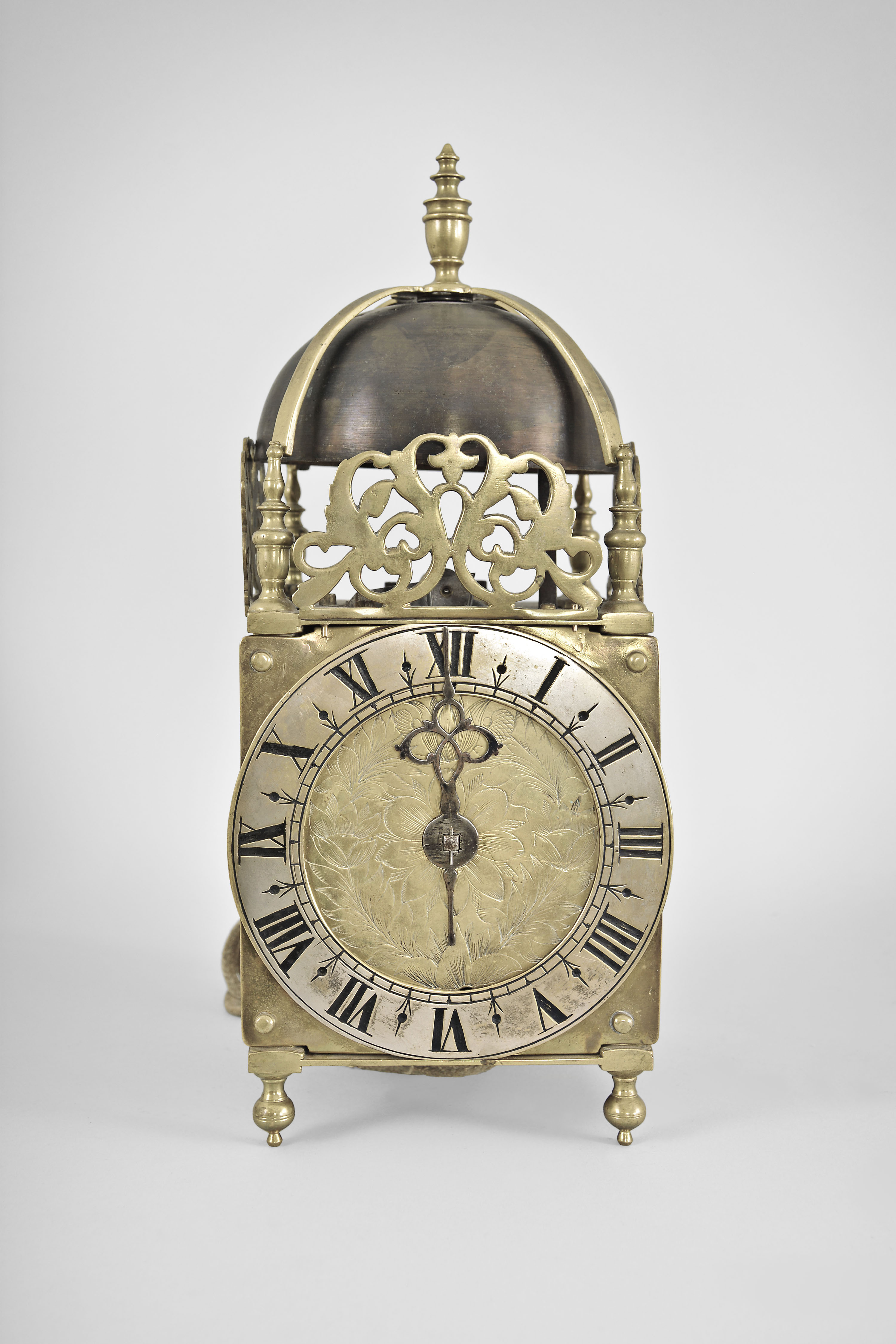 Probably London, England, an early 18th century striking lantern clock, the brass case with turned, tapered columns at the corners, with ball feet and slender urn finials, the top with arcaded brass gallery fixed to the top plate, front and sides with cast frets, the top with brass bell strap with finial and large bell, dial center with engraved foliage and tulips, the individual corners with a single, decorative button, silvered brass chapter ring with roman numerals and single steel hand, movement with recoil escapement, seconds pendulum, and count wheel striking, the case bottom with crude, cryptic engraving, and possibly dated 1717 — Antique Clocks Price Guide