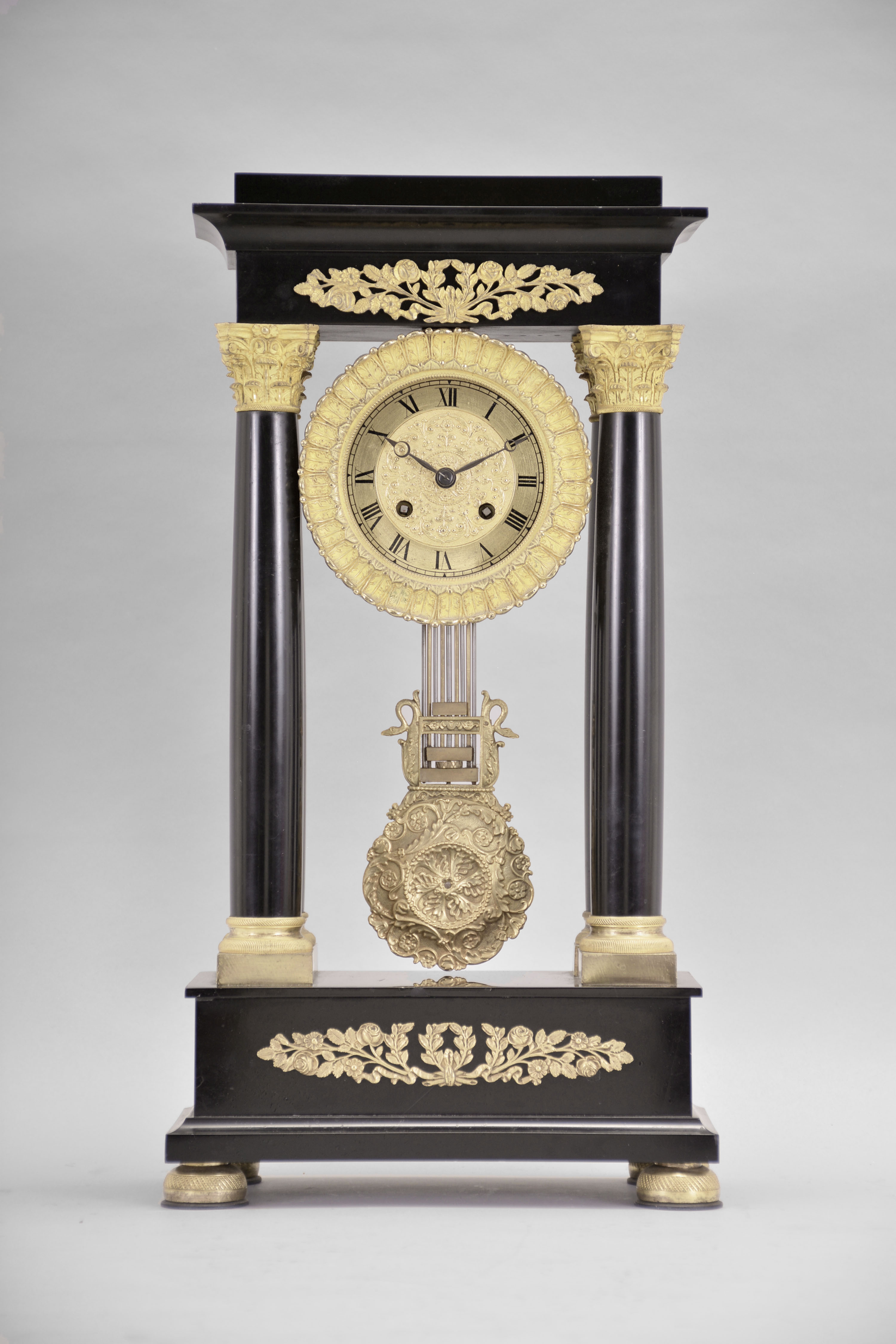 France, Empire portico clock, 8 day, time and strike, spring driven pendule de Paris movement in a polished black slate case on cast brass feet, the base and top with applied gilt rose ornaments, four Corinthian columns with gilt bases and capitals, the gilt bezel with repeating acanthus leaf design, gilt chapter ring with Roman numerals, cast dial center and gridiron pendulum both with floral designs, blued steel Breguet hands. — Antique Clocks Price Guide