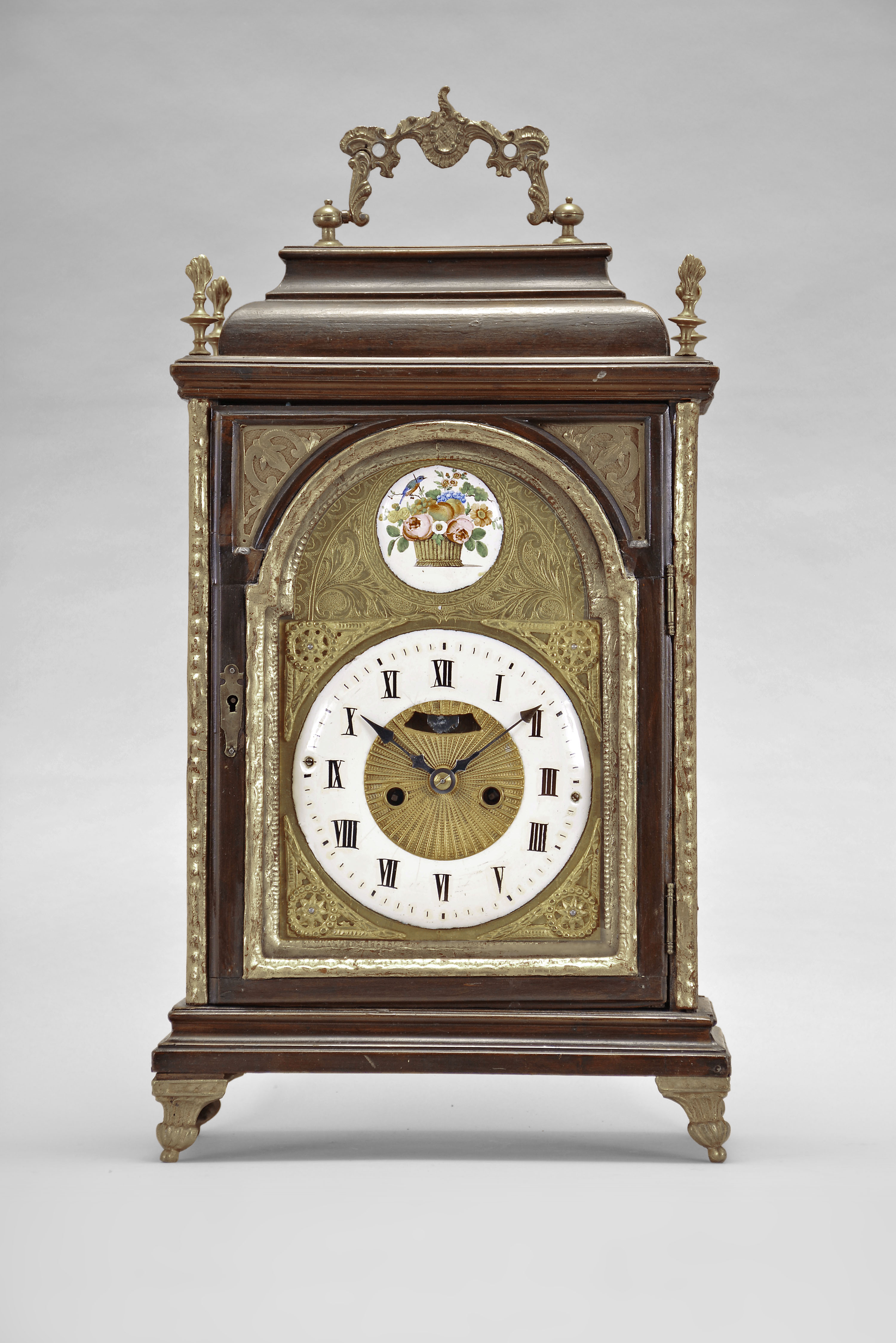 Austria, a late 18th century bracket clock, the molded base with cast, gilt feet, faux arch door with fabric backed frets at the upper corners, and caddy top, surrounded by four finials and with carrying handle, engraved brass dial with roman numeral, white enamel chapter ring, surrounded by cast spandrels, blued steel hands, patterned center with aperture for observation of mock pendulum, and boss in the arch with polychrome enamel scene with bird and basket of flowers, two train, two day movement with crown wheel escapement and rack striking, sounding the hours on a bell — Antique Clocks Price Guide