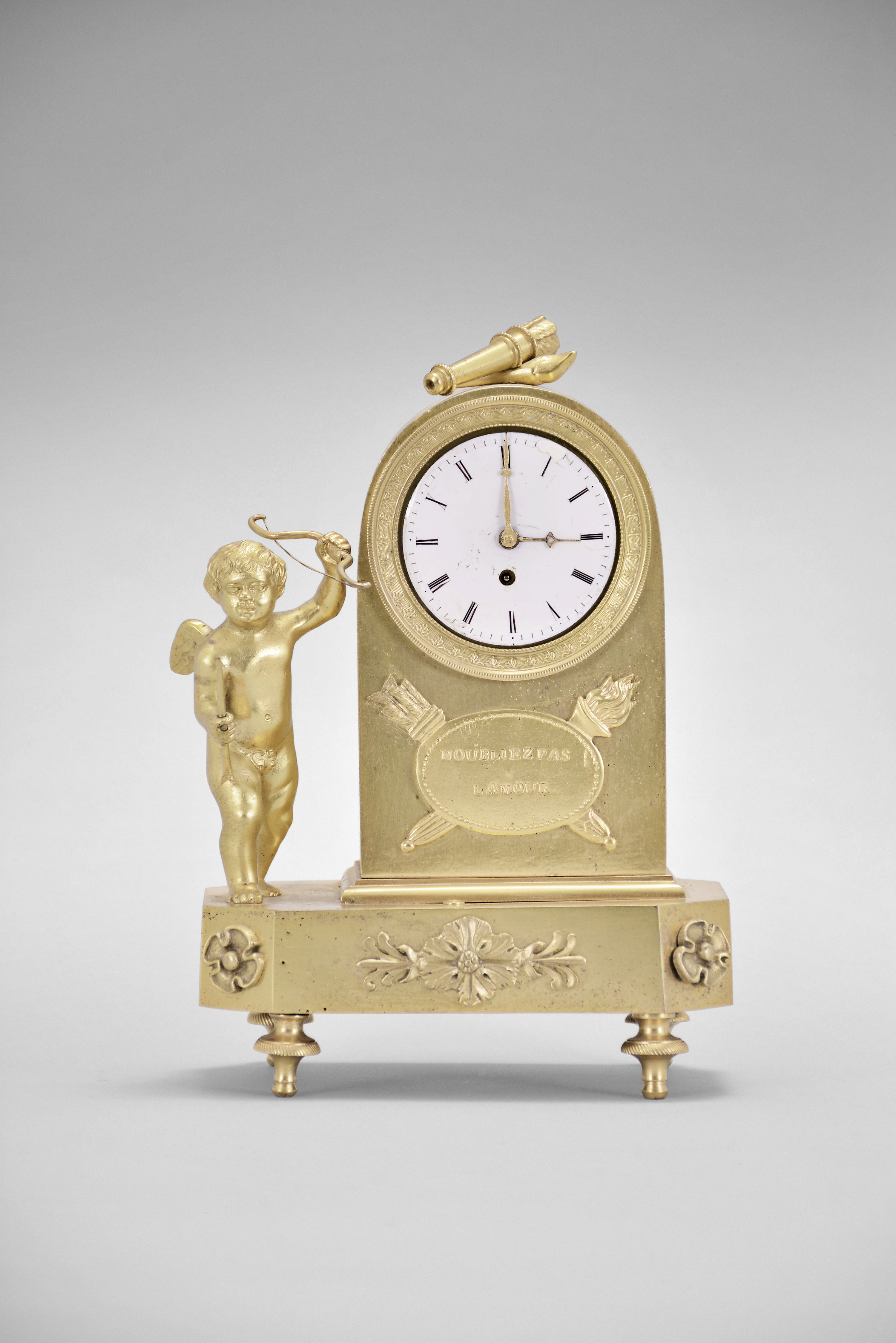 France, a small figural gilt bronze timepiece, the rectangular base with canted corners, toupie feet, with applied foliate ornament, and supporting a domed clock case with flanking figure of cupid, case top with torch and quiver, front with palmette ornamented bezel above an oval plaque before crossed torch and quiver, the plaque marked "N'oubliez Pas L'aomur", Roman numeral white enamel dial with gilt hands, and 30 hour verge fusee movement signed "Romilly Paris " — Antique Clocks Price Guide