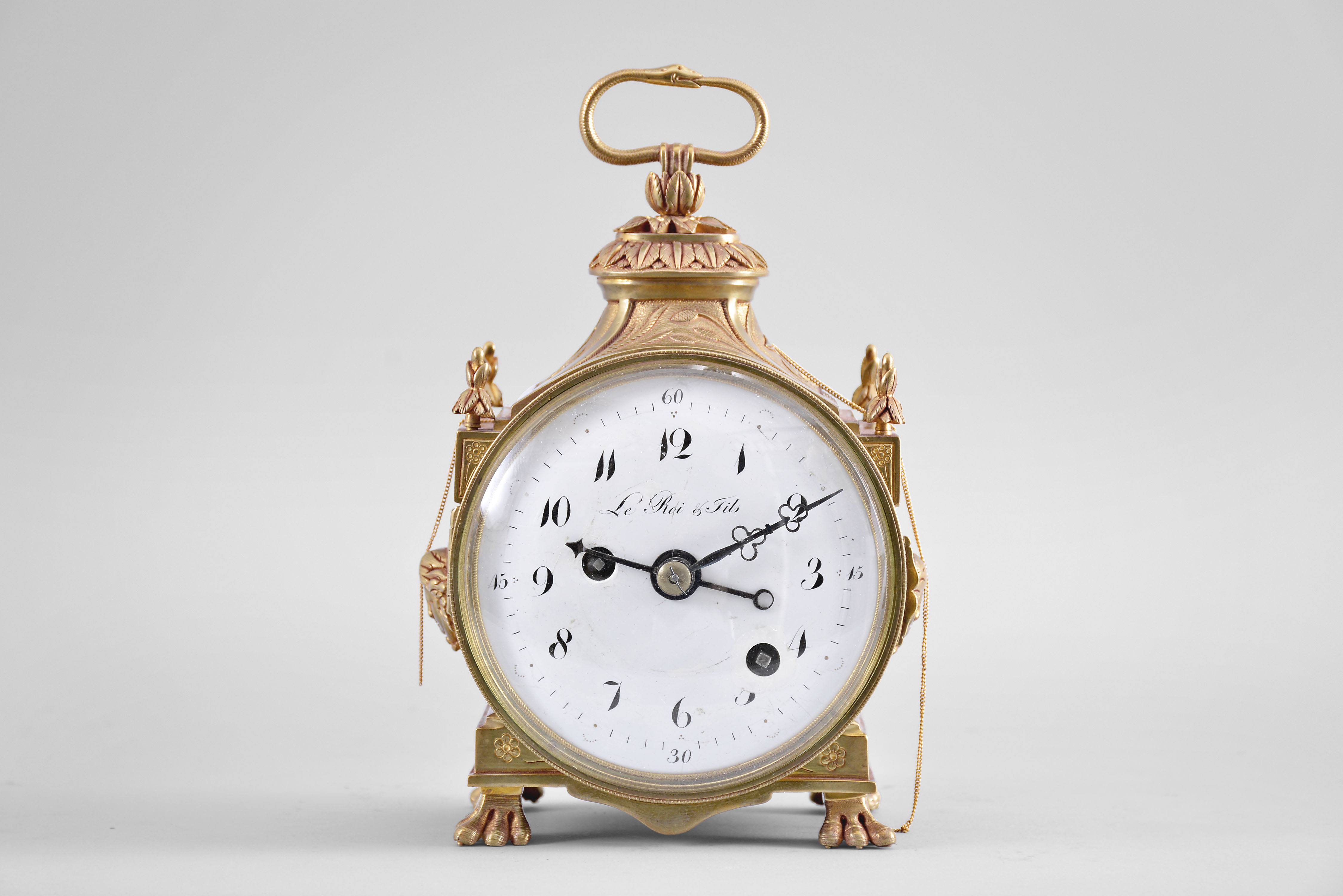 Le Roi &amp; Fils, Paris, a late 18th century grand sonnerie striking Pendule d'Officier, the round gilt bronze case with stepped, rectangular base on paw feet, the sides with applied Medusa masks below a wheat ear ornamented frieze and foliate finials at the corners, and with incurved top, surmounted by an oval, foliate ornament with folding ouroboros handle, hinged brass bezel with convex beveled glass, Arabic numeral white enamel dial, blued steel hands, 30 hour, grand sonnerie striking and repeating movement with alarm, time train with fusee, strike train with barrel, the alarm wound by pulling a fine chain at the left side of the case, the repeating activated through a chain at the right, strike work mounted to back plate, and with verge escapement, the plain three arm balance visible beneath the pierced balance bridge, mounted to the back plate, the back plate stamped "SRF " — Antique Clocks Price Guide