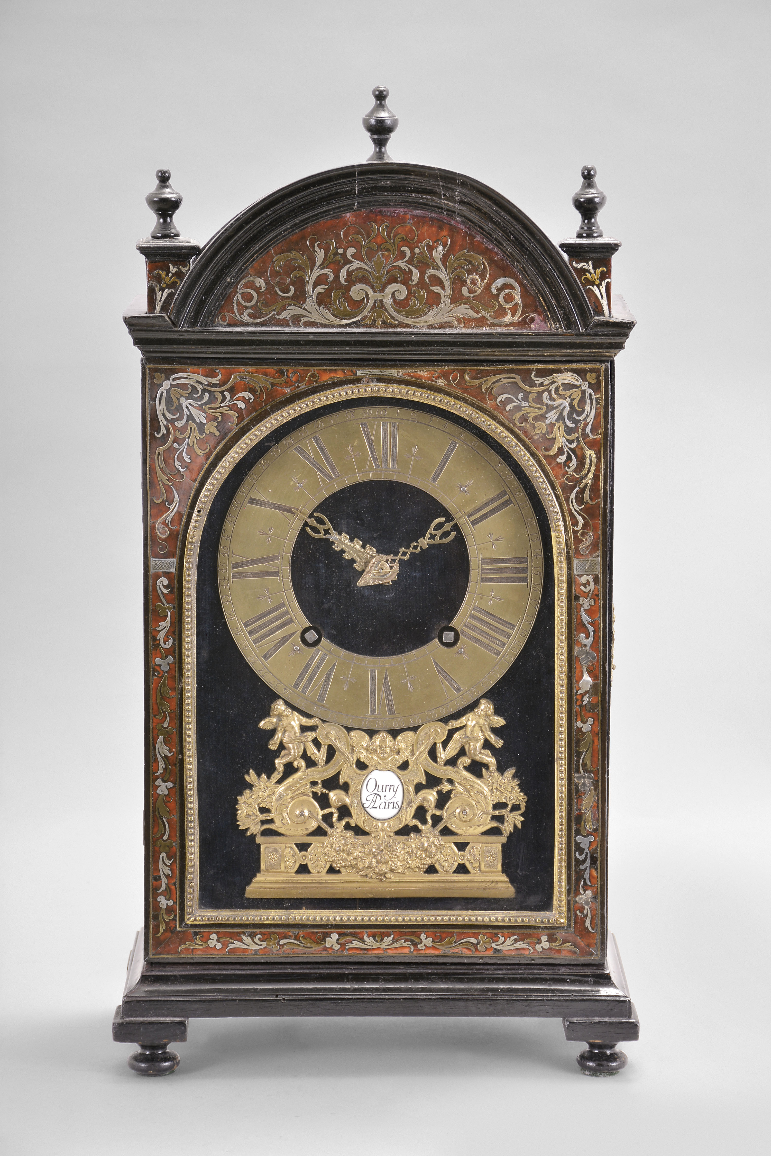Ourry, a Paris, an ebonized Pendule Religieuse with Boulle work front, the case with molded base and turned feet, case sides with tombstone arch windows, case front with tortoise shell veneers and foliate form brass and pewter inlays, door glass with cast, arched, beaded trim, top with arched, molded tympanum, flanking plinths, and three finials, dial plate lined with deep blue velvet, engraved brass Roman numeral dial with Arabic minutes, fancy cast brass hands, and with cast, gilt brass mount below 6:00, consisting of a balustrade on a molded base supporting foliate ornamented S scrolls, and surmounted by putti, with central enamel cartouche marked "Ourry A Paris", rectangular movement with five baluster form pillars and large barrels, recoil escapement, and counting the hours on a bell, back plate also signed — Antique Clocks Price Guide